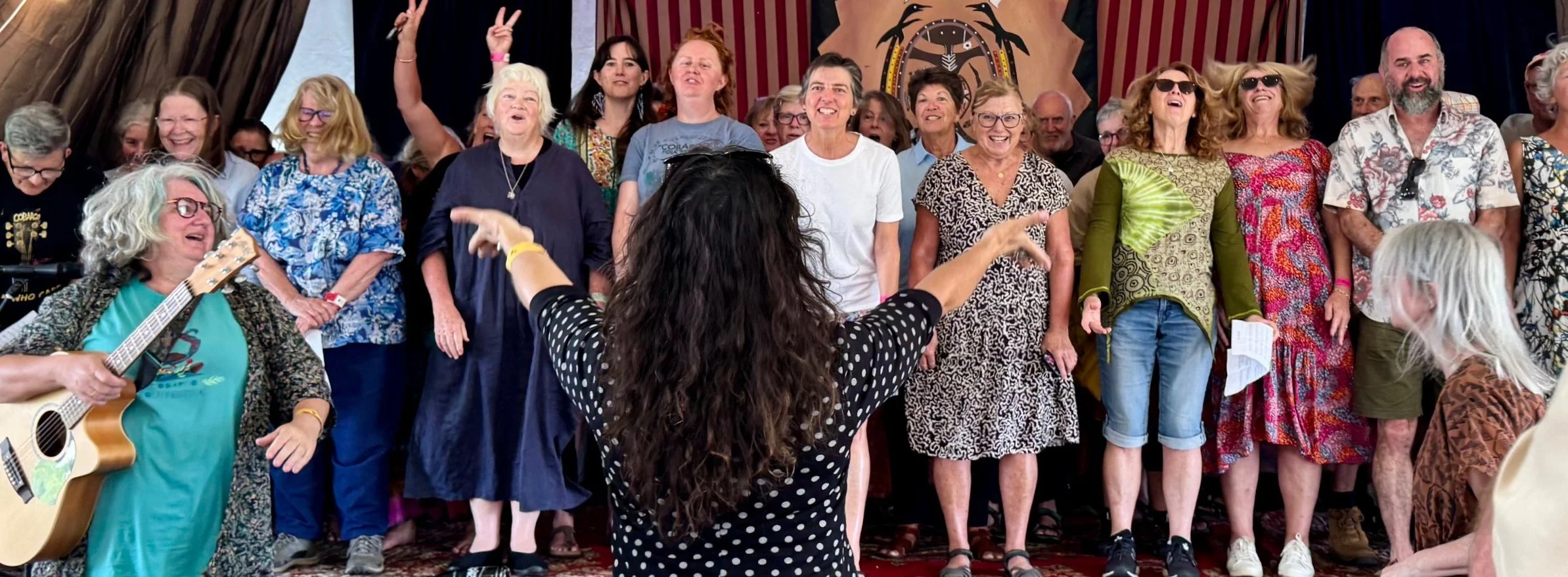 A group of people singing on stage with a woman conducting, some wearing sunglasses, with a woman playing guitar in the foreground.