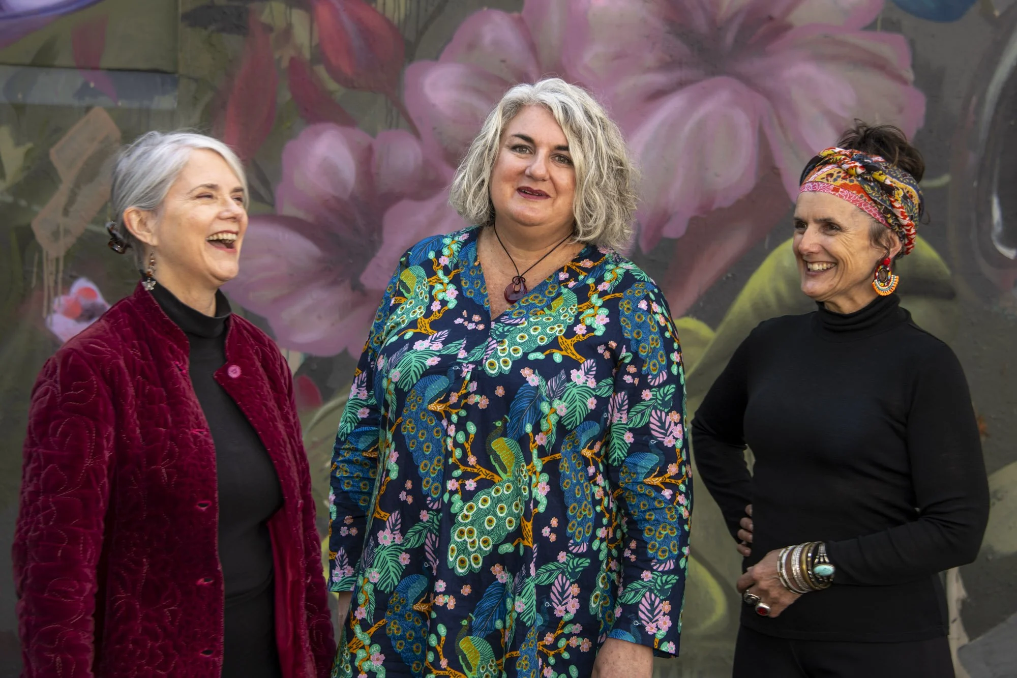 Three women standing together outdoors in front of a large floral mural. The woman on the left has short gray hair, is wearing a red quilted jacket and black turtleneck, and is laughing. The woman in the middle has shoulder-length curly gray hair, is wearing a vibrant, floral-patterned jacket, and is smiling softly. The woman on the right has dark hair under a colorful headscarf, is wearing a black top and large colorful earrings, and is smiling. All appear to be enjoying a moment together.