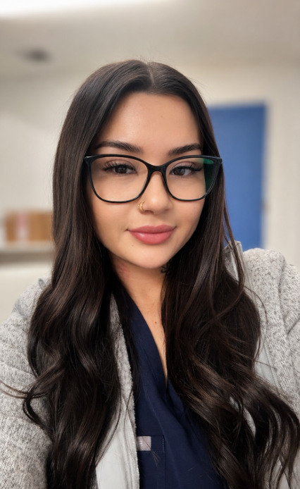 A young woman with long dark hair, glasses, and a nose ring taking a selfie indoors.