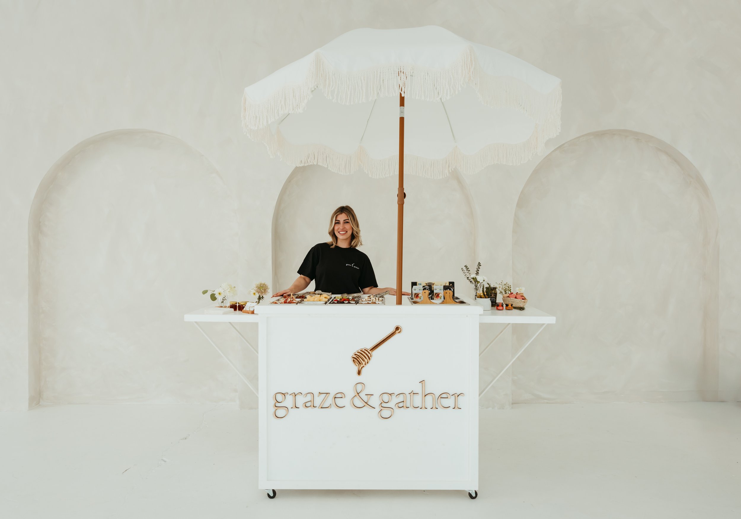 Person standing behind a white cart with "graze & gather" written on it, featuring food displays, under a white fringed umbrella in a minimalist setting. - graze & gather | Luxury Charcuterie in Jacksonville, Florida