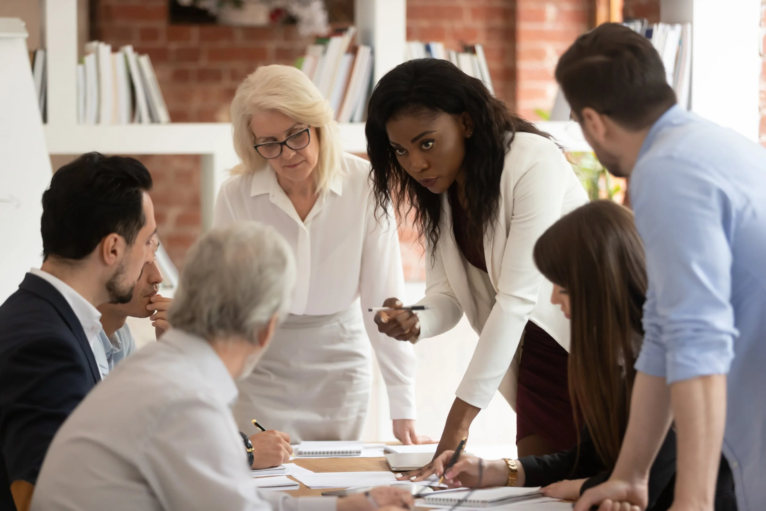 Six people gathered around a table in a meeting, engaged in discussion. A woman with dark hair is standing, leaning over the table, holding a pen and talking. Others are seated, listening and taking notes. The setting is an office with a bookshelf and brick wall in the background.