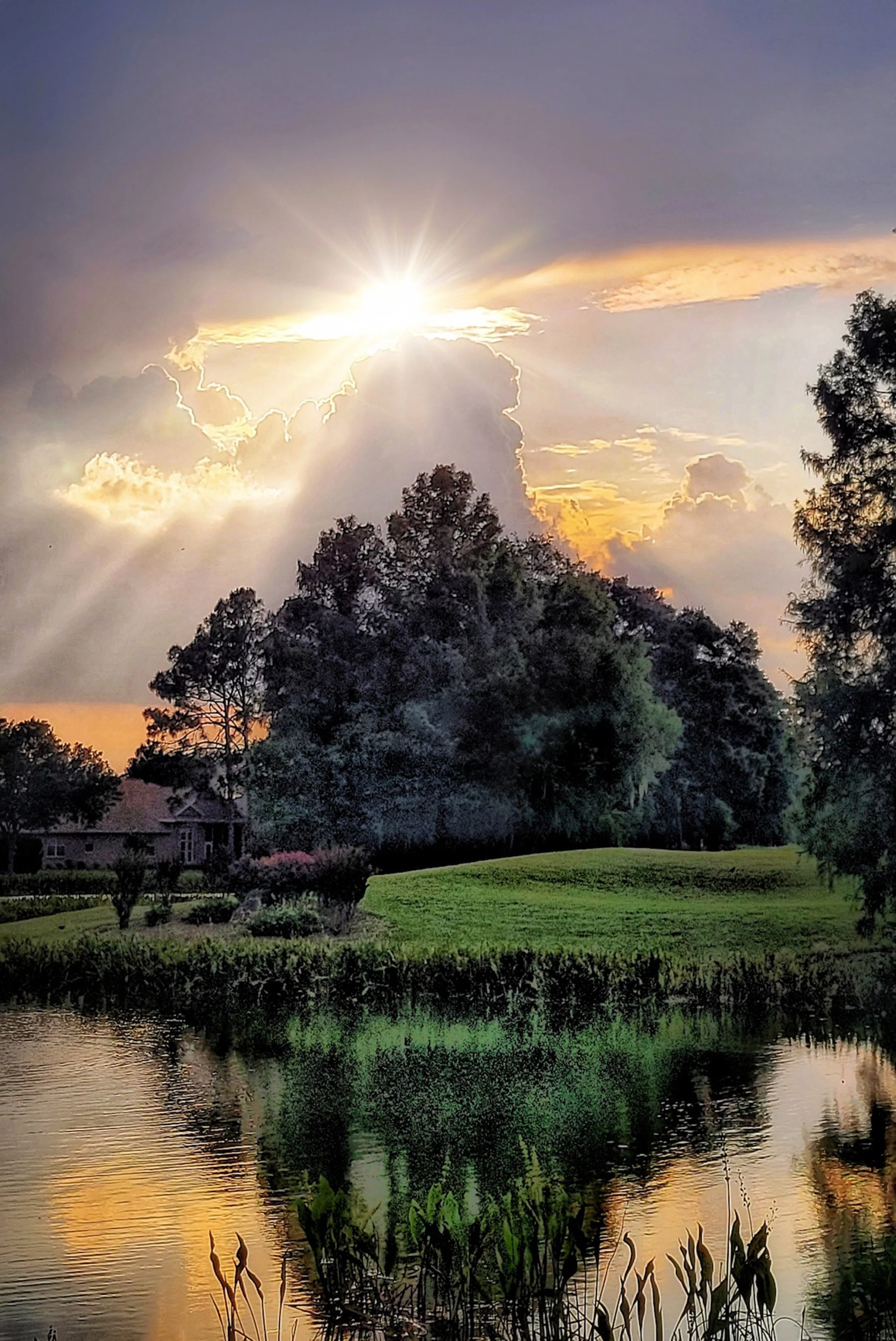Sunset over a landscape with trees, a house, a pond, and colorful clouds in the sky.
