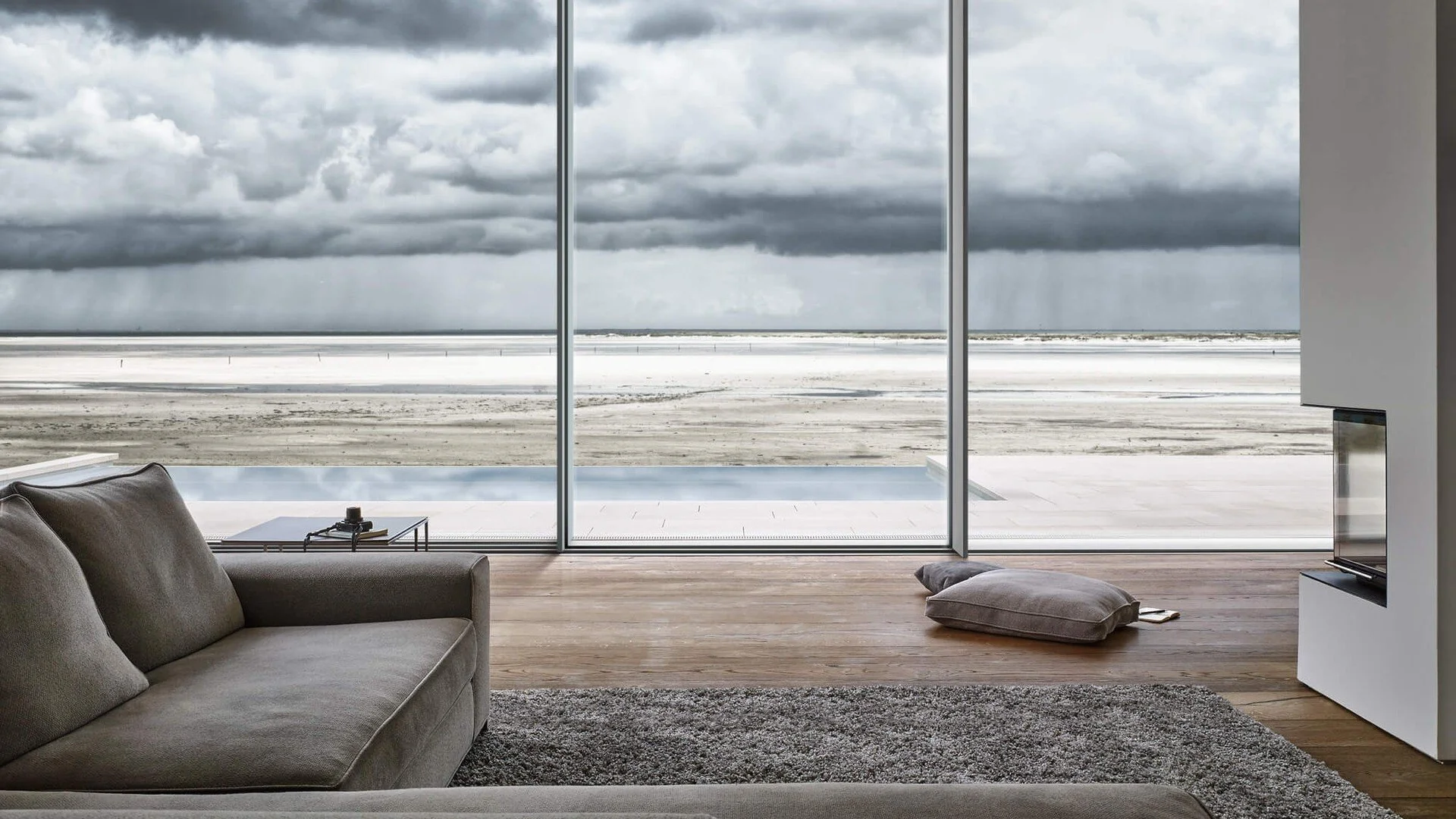 Modern living room with large glass window overlooking a snowy landscape and dark cloudy sky, featuring a gray sofa, a small black side table, a gray cushion on the floor, and a TV on a white stand.