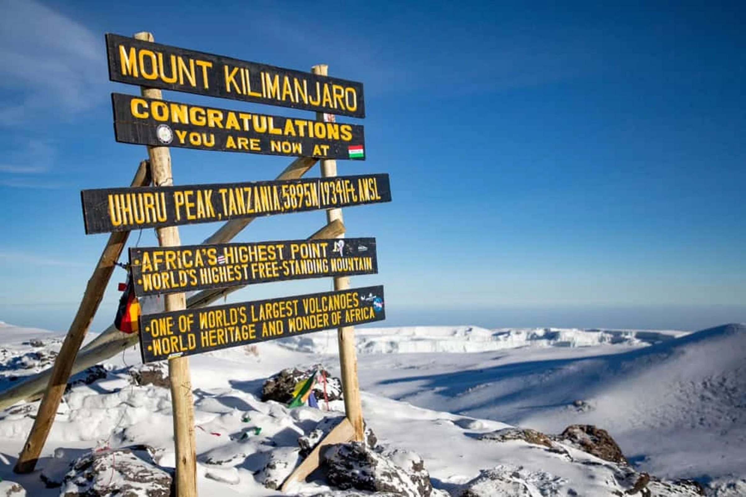 Sign at Uhuru Peak, Tanzania, indicating it is the highest point in Africa at 5895 meters (19341 feet) above sea level, celebrating reaching Mount Kilimanjaro, the world's highest free-standing mountain and home to one of the world's largest volcanoes. The snow-covered landscape and clear blue sky are visible in the background.