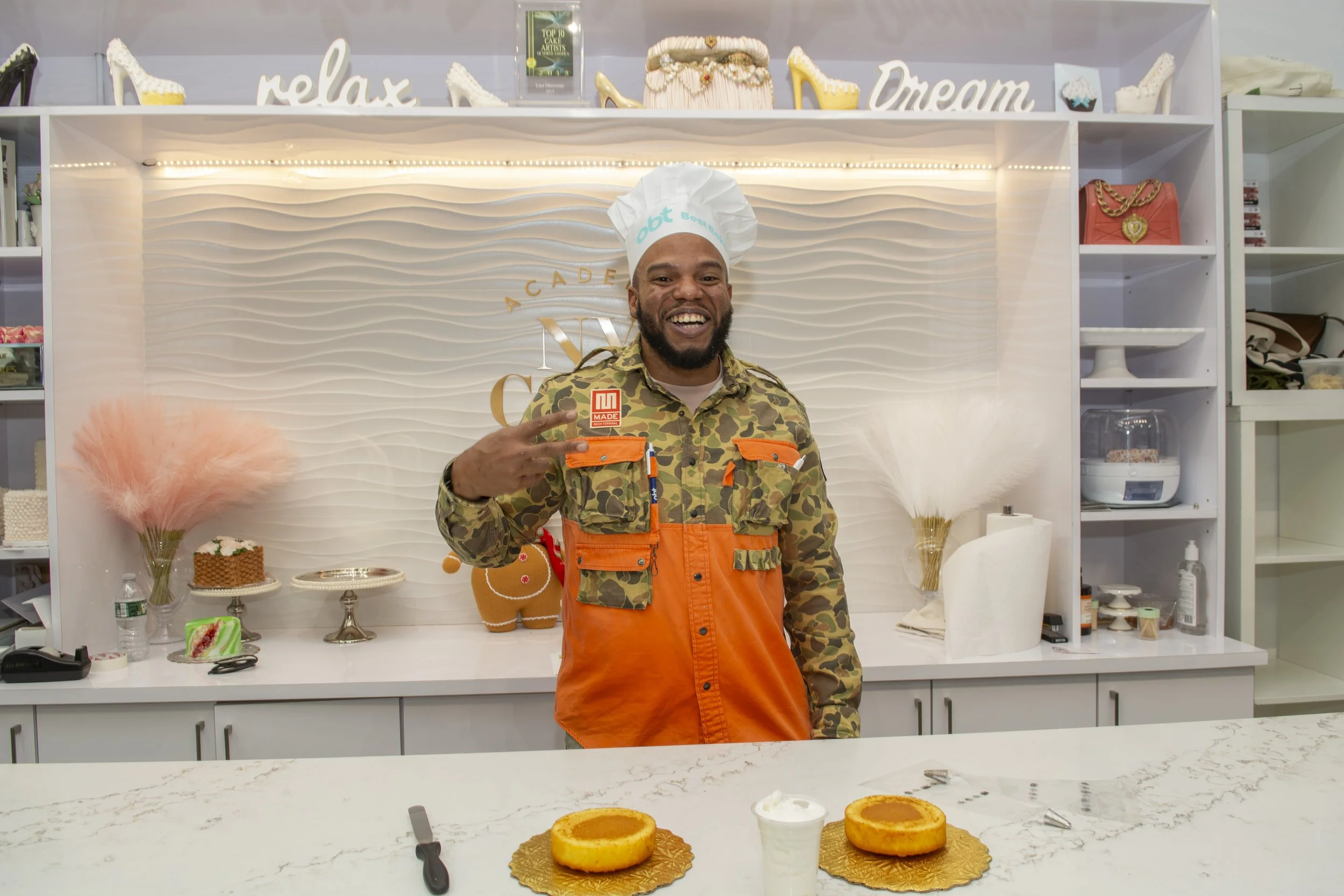 Kareem Youngblood smiling in NY Cake kitchen with two round cakes on gold doilies and a glass of white beverage in front of him, wearing a camouflage and orange outfit, and a chef hat with the word 'Bert' embroidered.