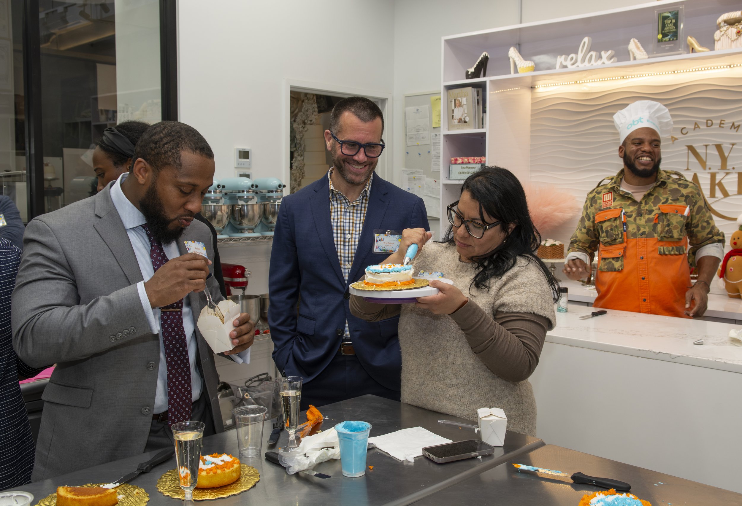 People celebrating a birthday with cake in a kitchen, including a woman cutting a cake with a blowtorch, and others watching and smiling.