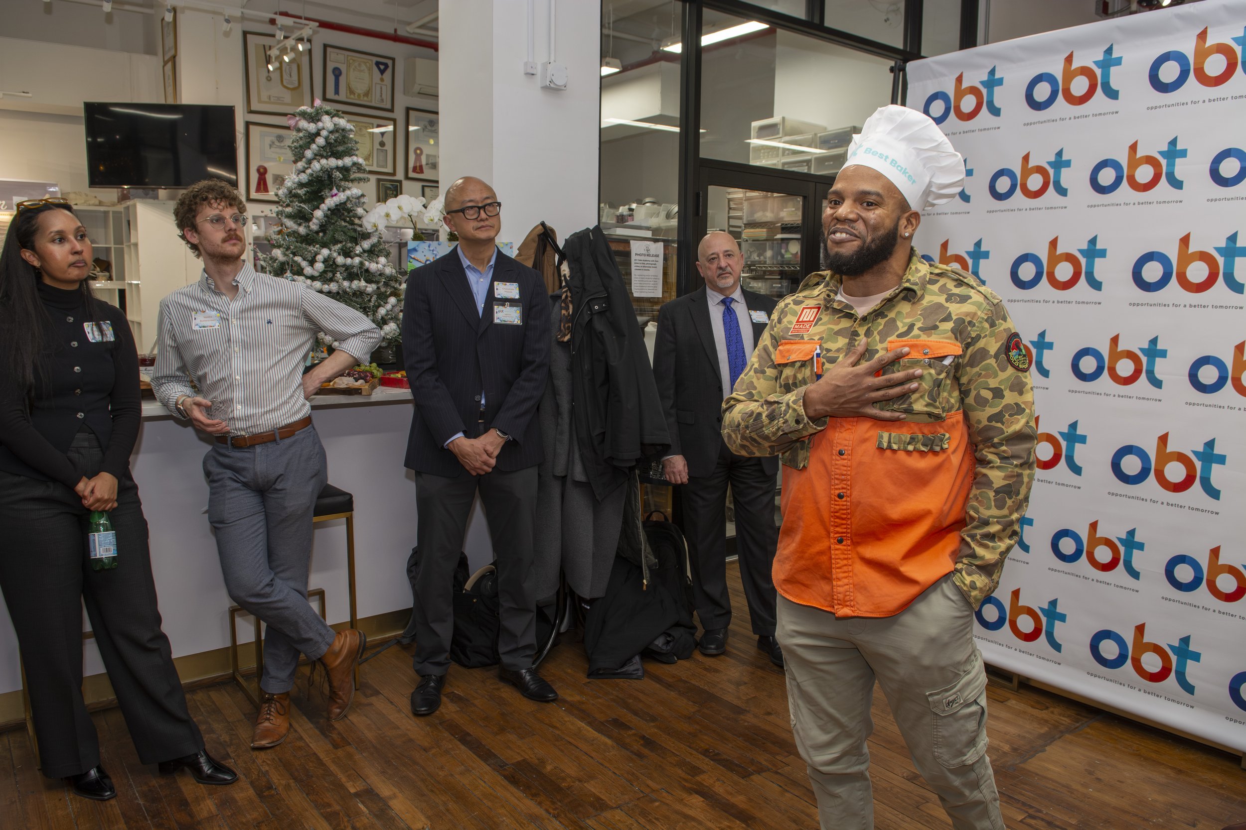 A man wearing a camouflage jacket, orange apron, and a chef's hat stands in front of a blue and orange 'obt' banner, with his hand over his chest, speaking or presenting to an audience of five people in a festive room decorated for Christmas.