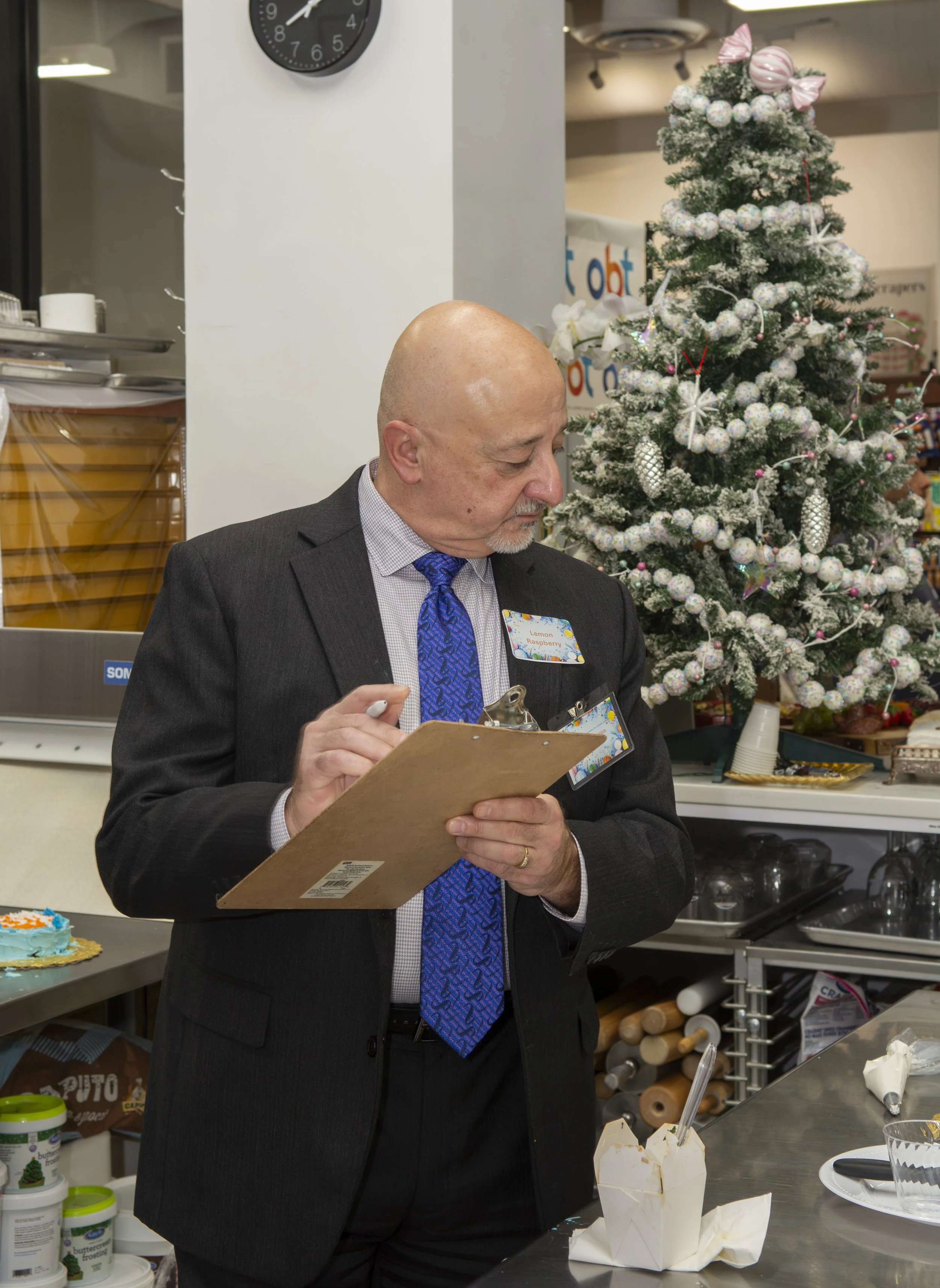 A man in a suit with a blue tie holding a clipboard and writing, standing next to a decorated Christmas tree in a store or restaurant setting.