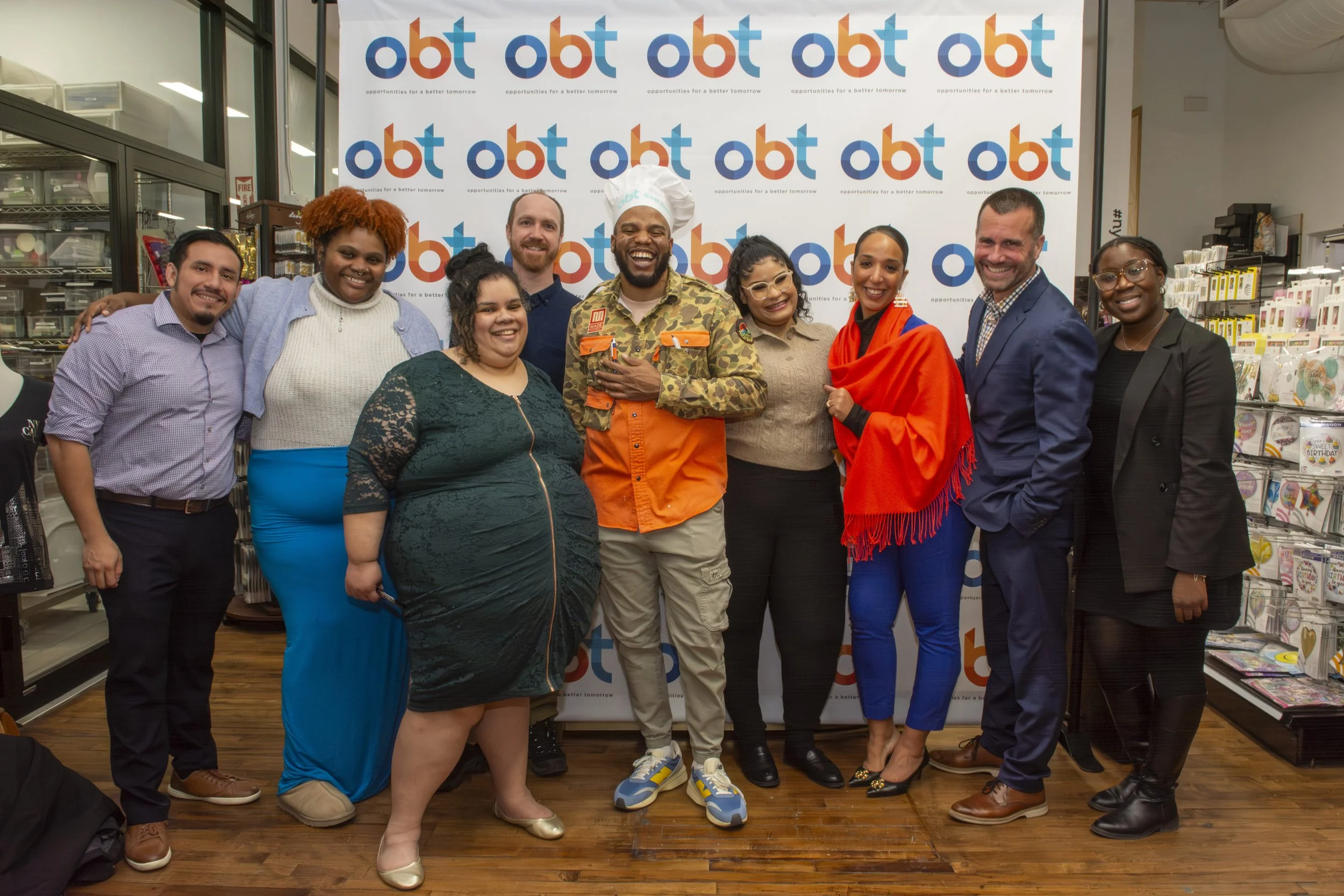 A diverse group of nine people, including men and women of different ages and ethnicities, standing together and smiling in front of a step-and-repeat banner with the logos 'OBT' and the tagline 'opportunities for a better tomorrow'. They are inside 