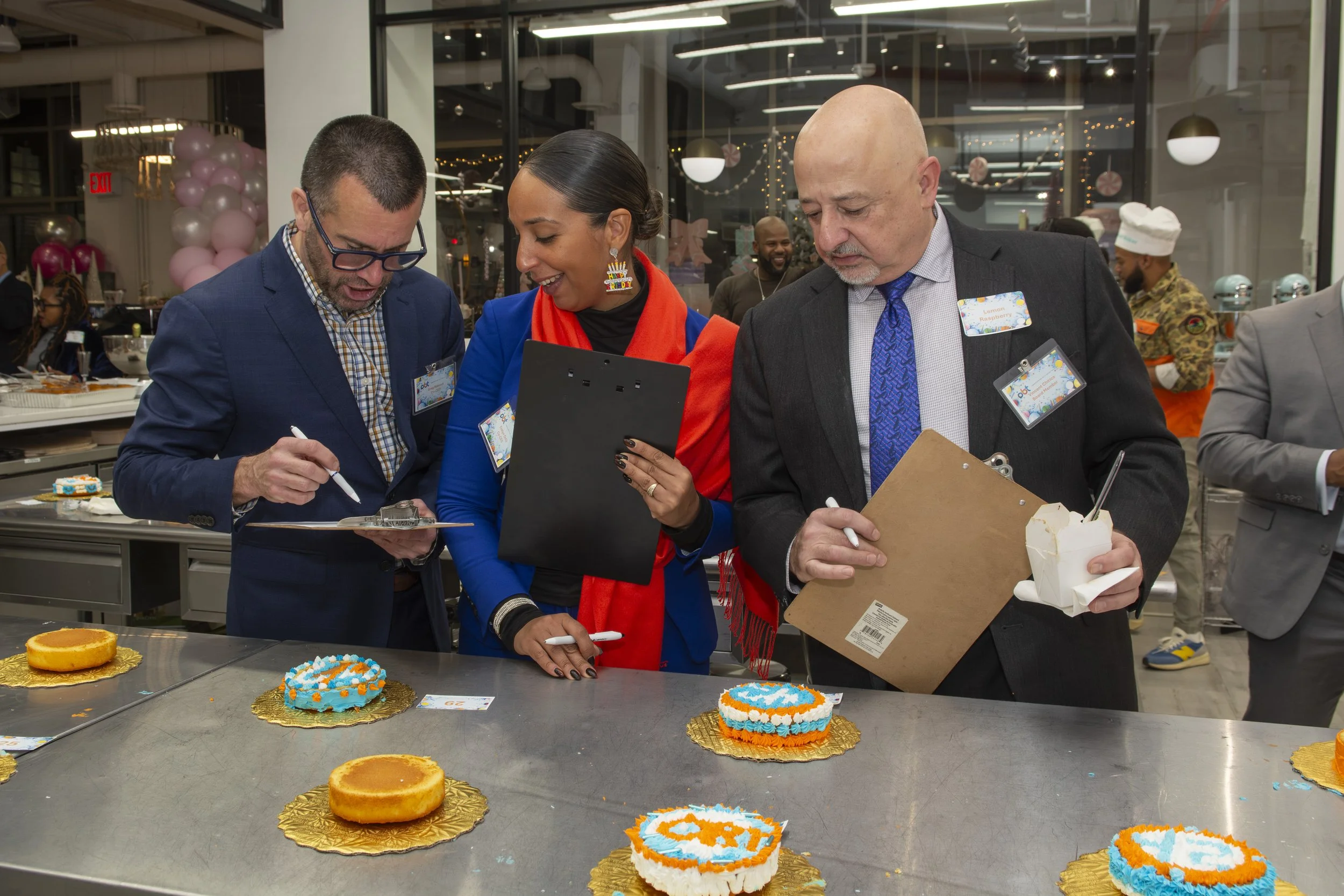 Group of three people in business attire at a celebration, examining decorated cookies on a table covered with gold foil, with balloons and festive decorations in the background.