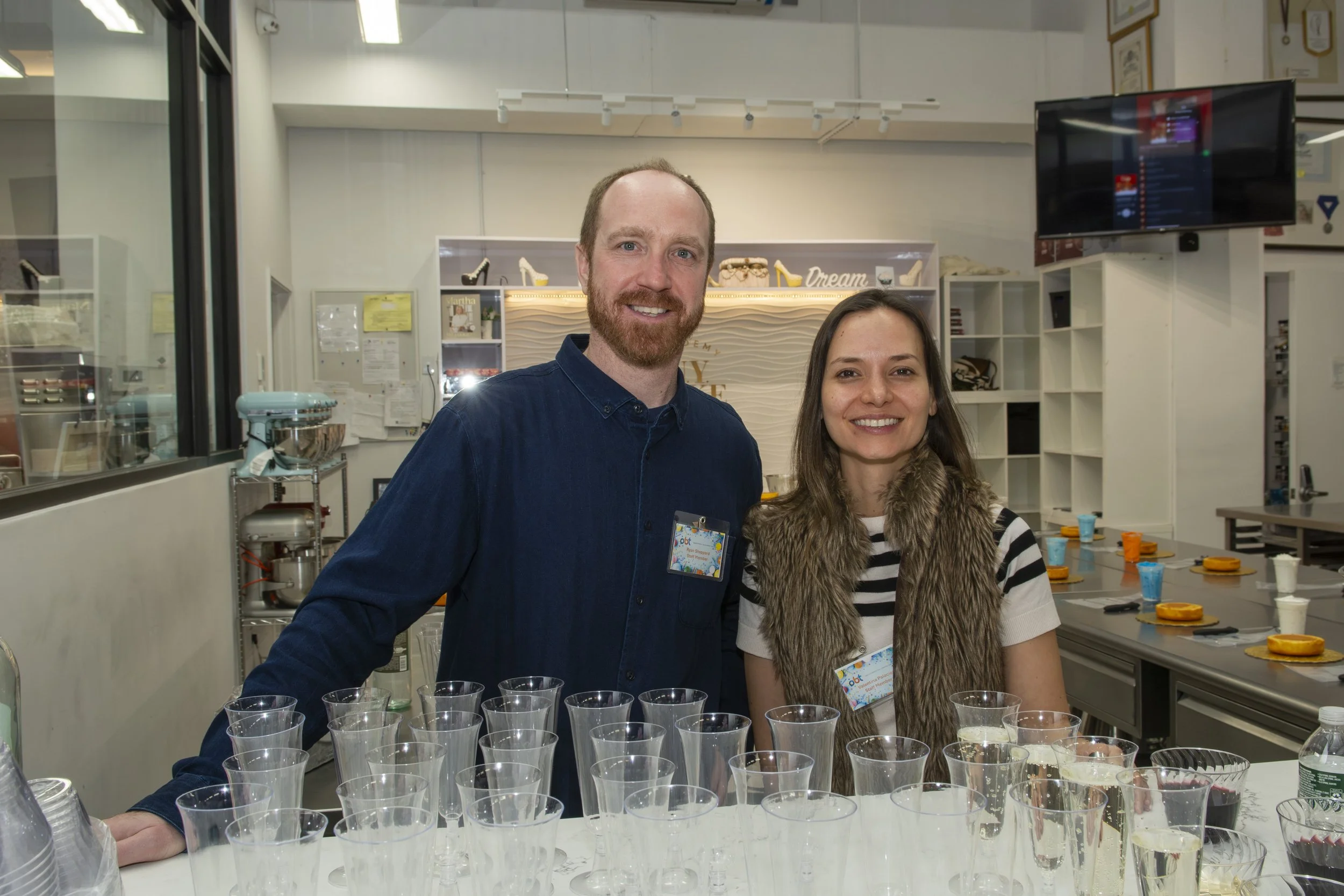 Two smiling people, a man and a woman, standing behind a counter with clear plastic cups and cups with orange and white lids, in a room with shelves and a large TV screen.