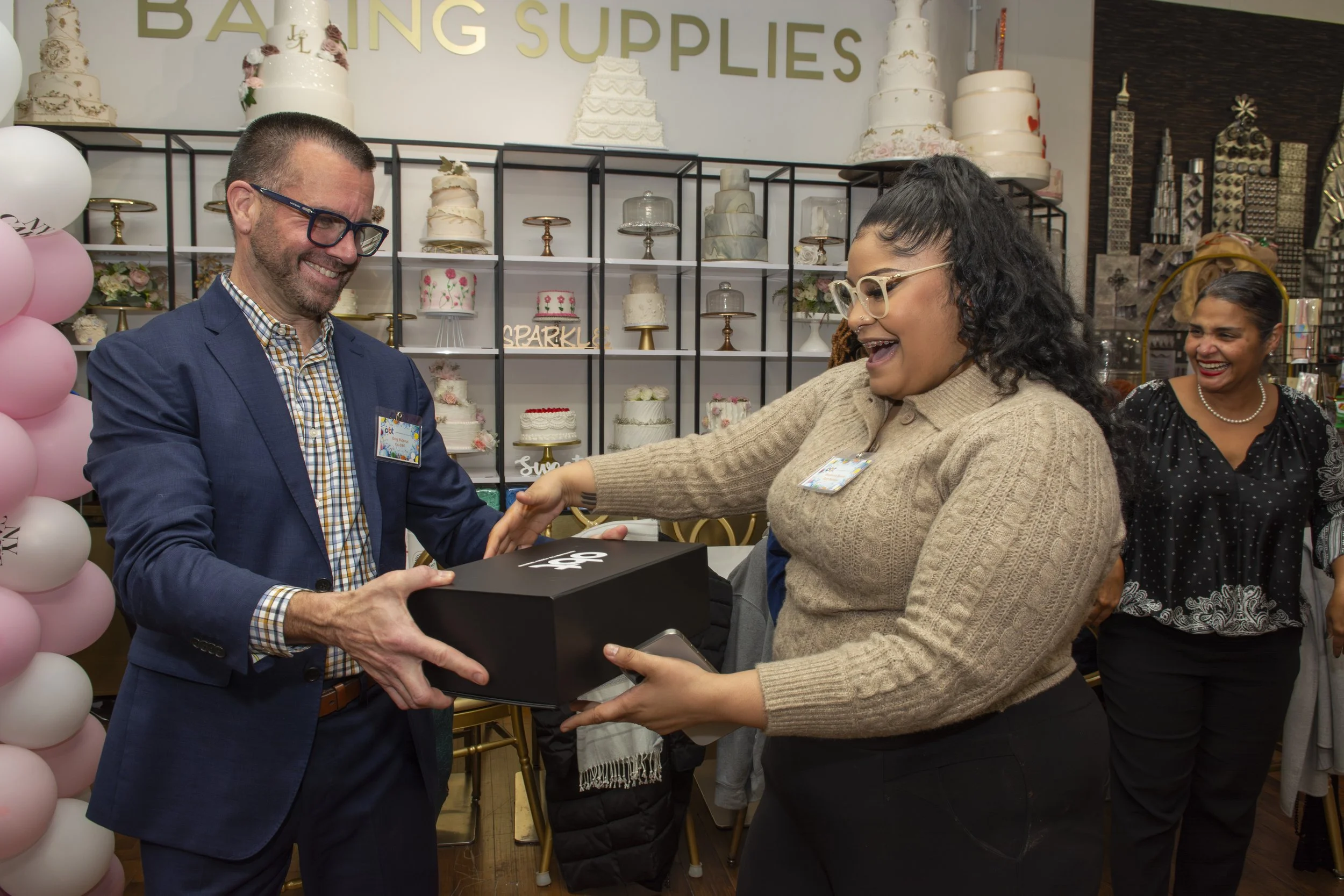 A woman in a beige sweater and glasses is receiving a black box gift from a man in a blue blazer at a cake shop. Another woman is smiling in the background. There are decorated cakes on shelves and pink and white balloons to the side.