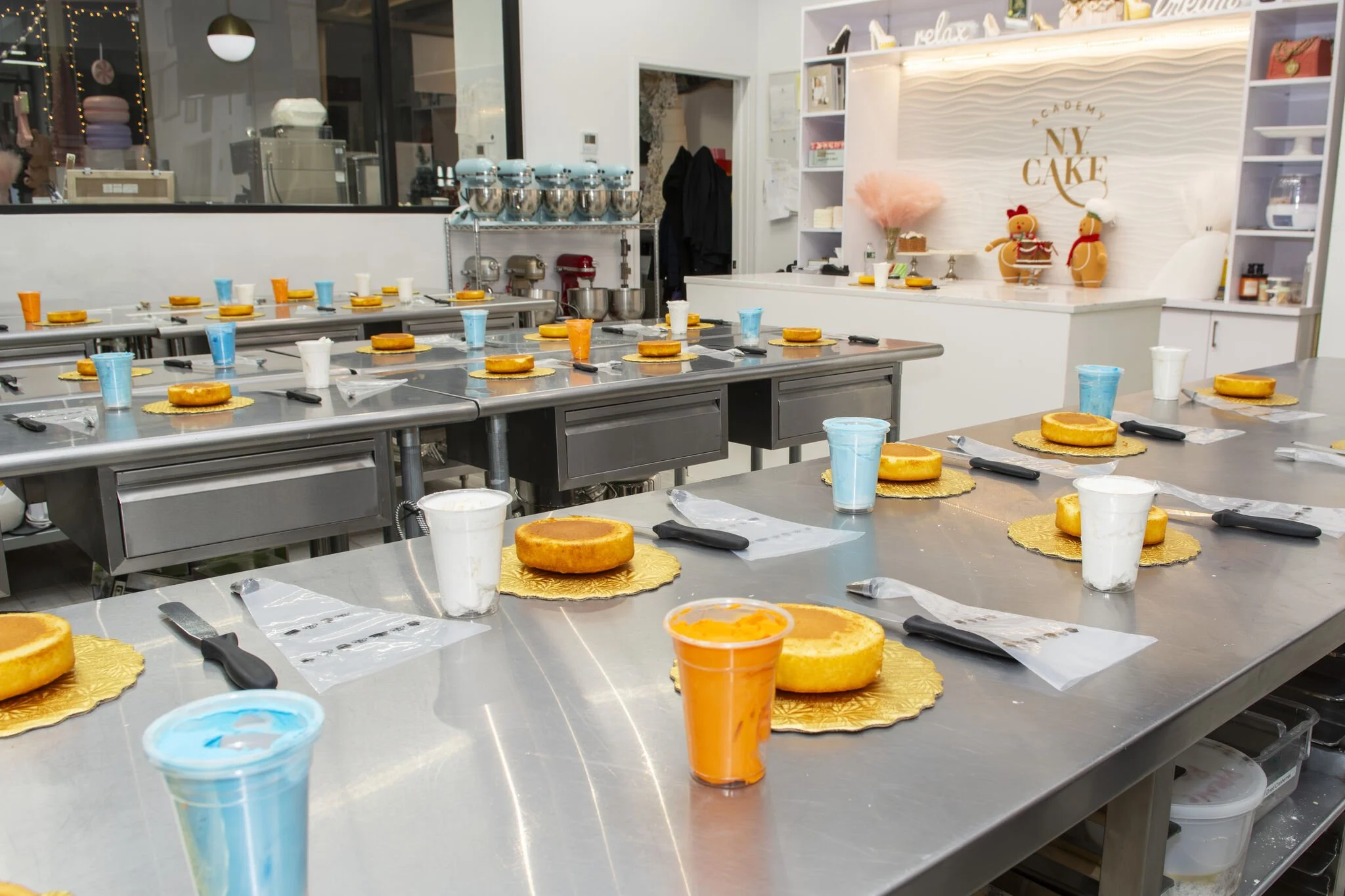 A cake decorating class setup with tables, each having a round cake on a gold doily, plastic cups filled with blue and white icing, and cake knives covered in plastic. In the background, there are metal mixing bowls, stand mixers, and a decorated wal