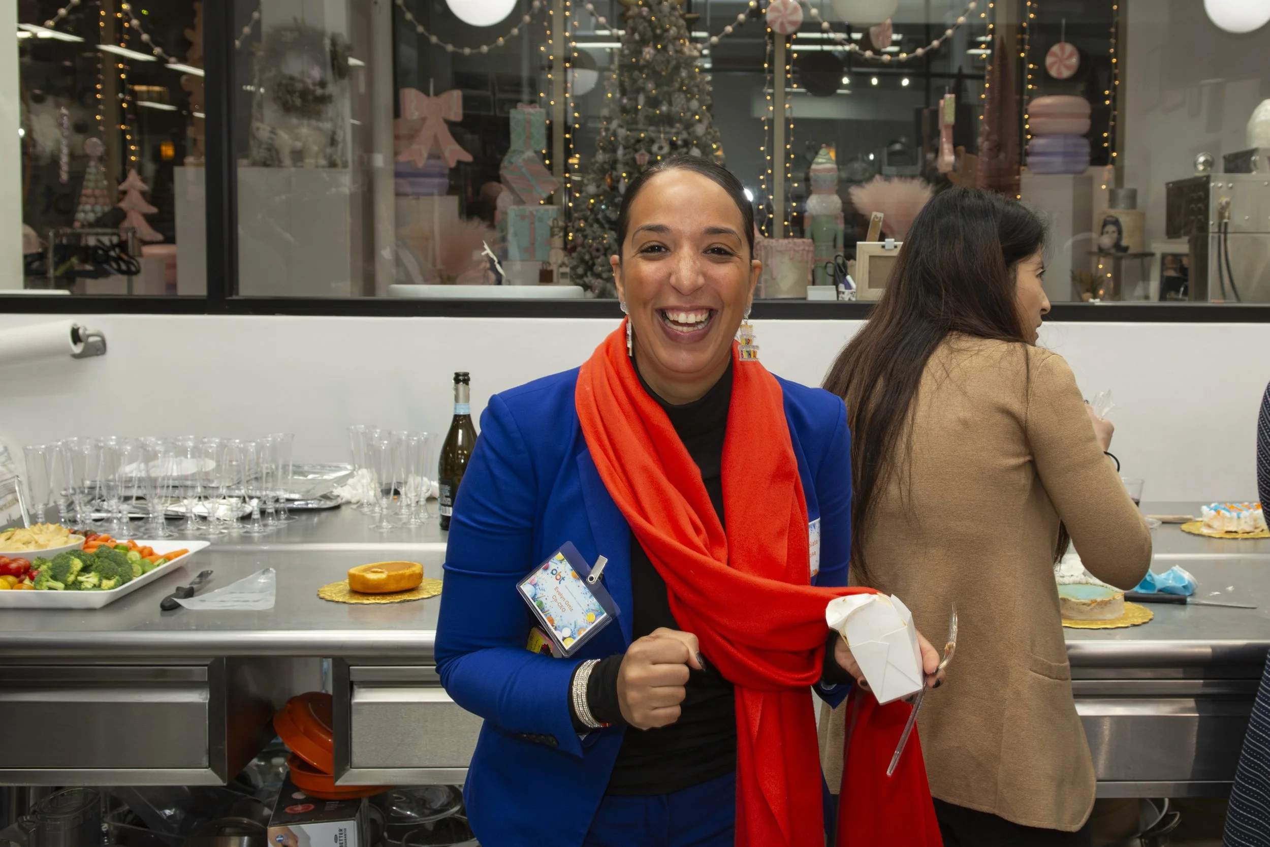 A woman wearing a blue blazer and orange scarf, smiling at the camera, standing in a festive setting with holiday decorations and a Christmas tree in the background.
