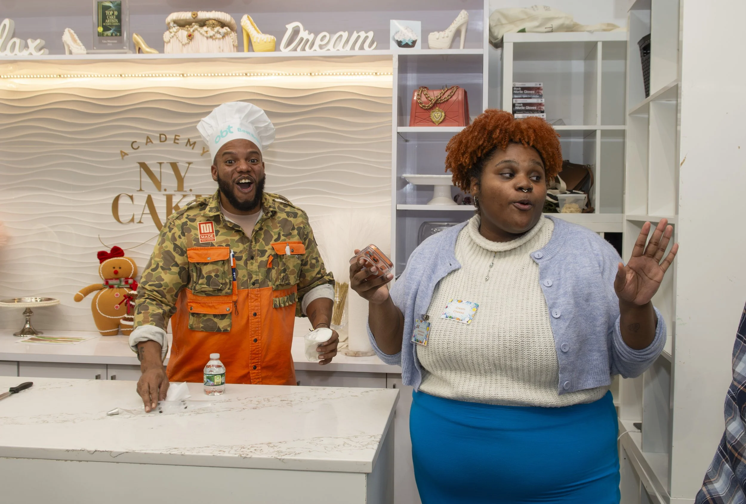 A man in a chef's hat and colorful apron stands behind a counter with a surprised expression, while a woman in a gray sweater and blue skirt gestures with her hand and appears to be speaking, inside a bakery or cake shop with decorative items on shel
