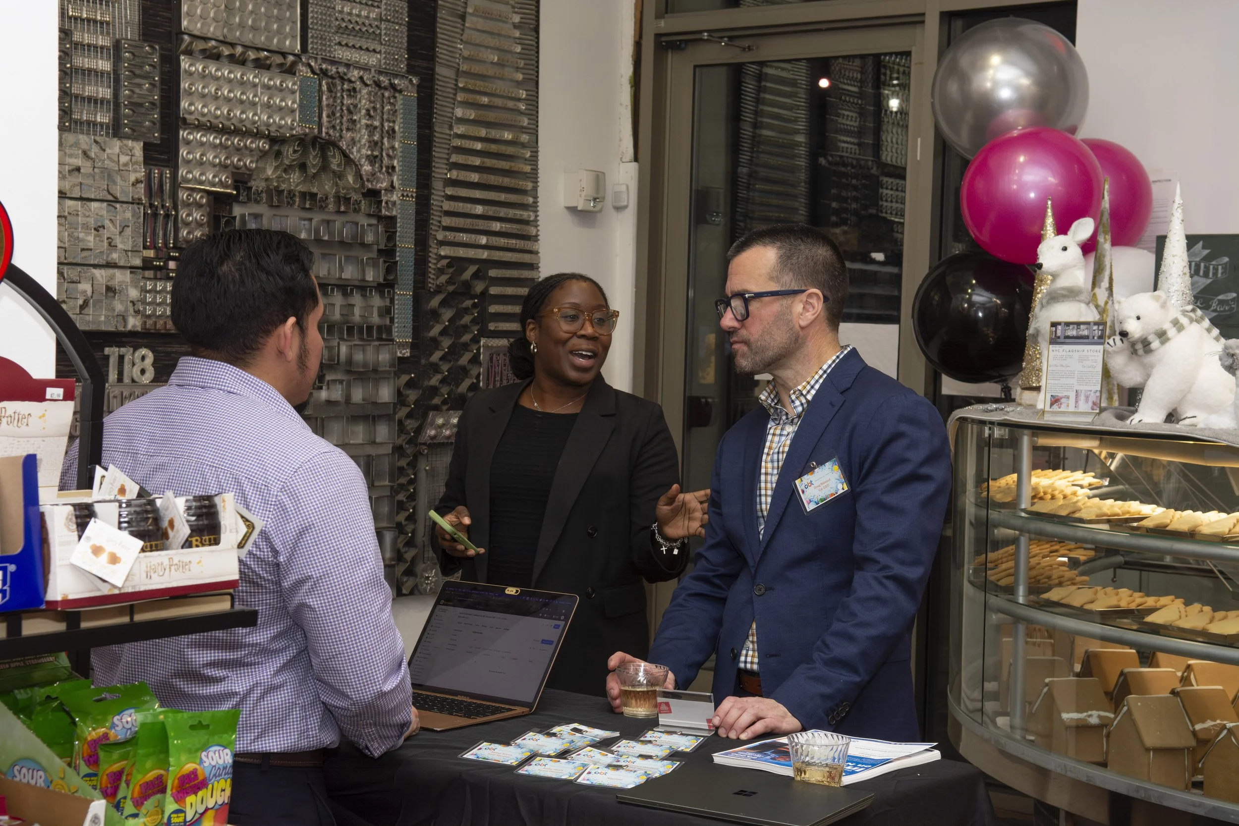 Three people having a conversation at a bakery or cafe. One is a woman with glasses and a black blazer, talking to two men in business attire. The man on the left has his back to the camera, and the man on the right is holding a drink. The scene incl
