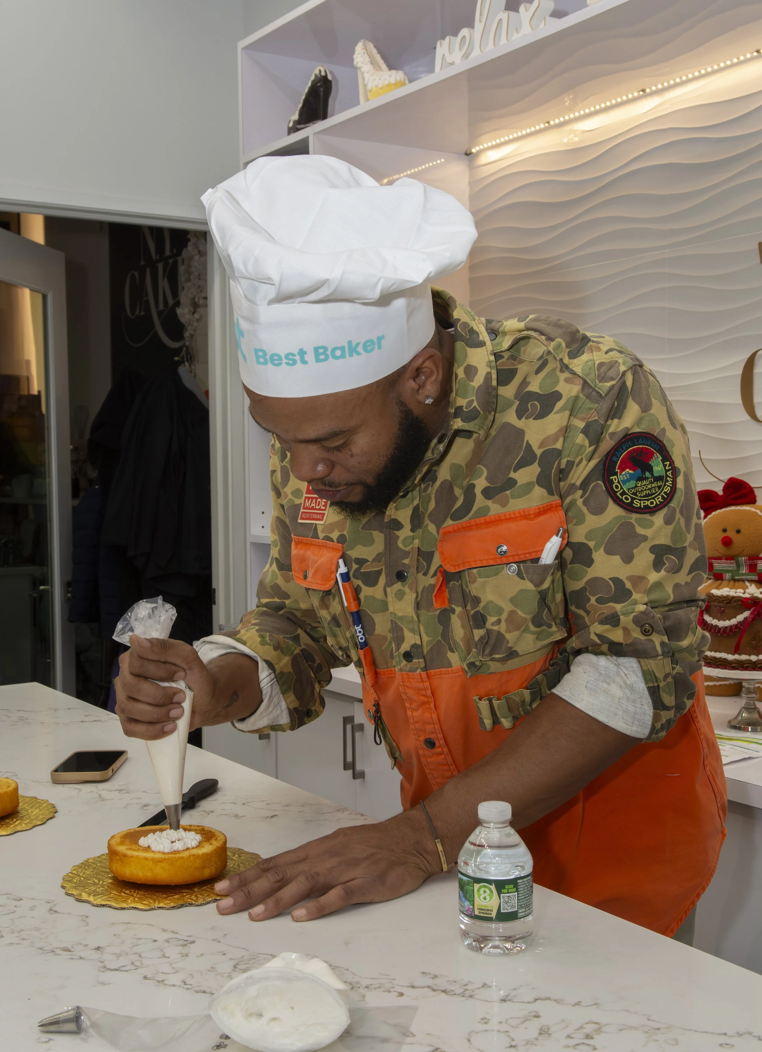A man wearing a chef's hat with 'Best Baker' written on it, a camouflage shirt with orange accents, and an orange apron, is decorating a cake with white icing in a kitchen decorated for Christmas.