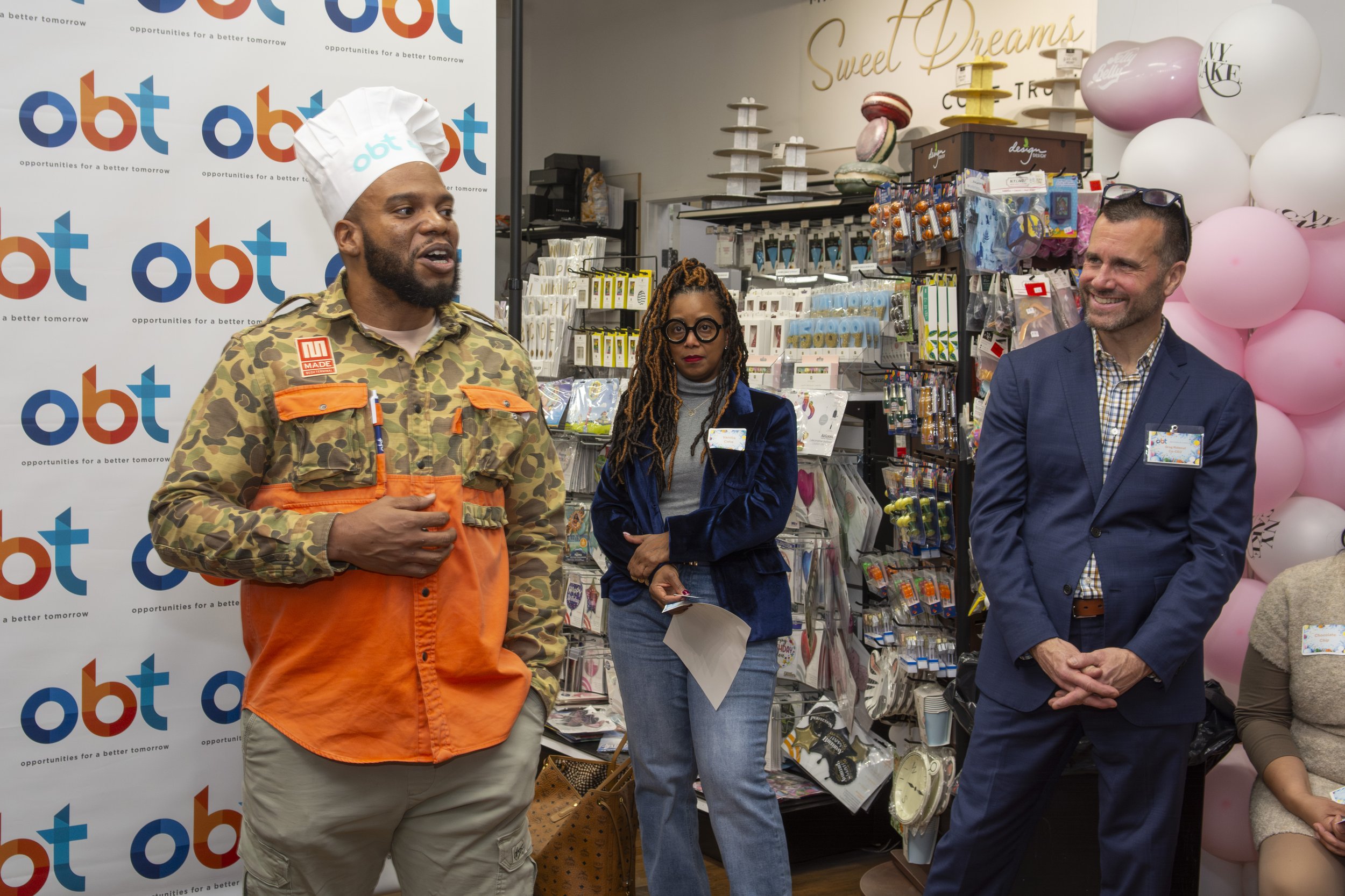 Three people standing in front of a gallery wall in a store, with one man speaking and two others listening, surrounded by store merchandise and pink balloons.