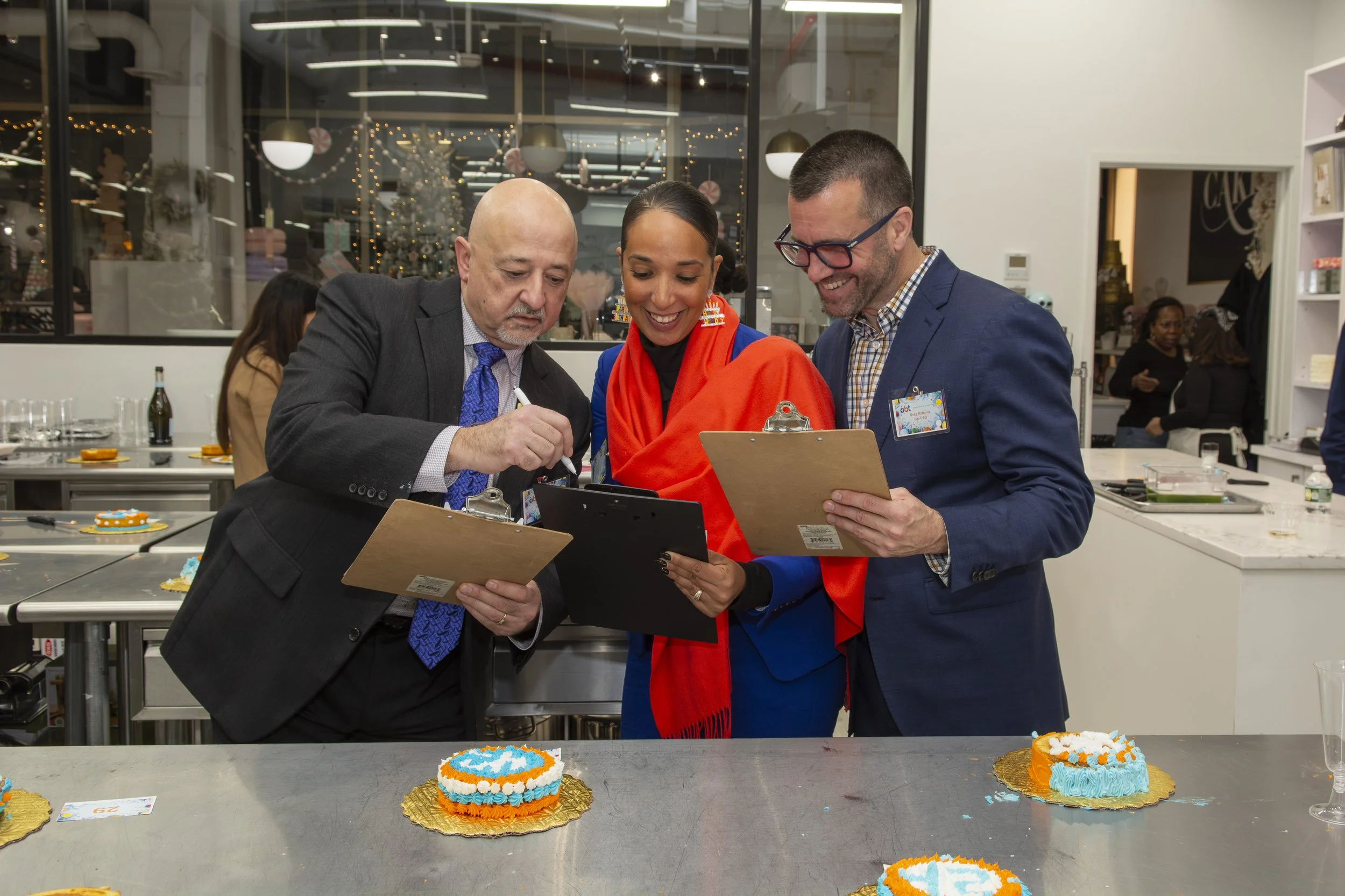 Three people in business attire smiling and looking at clipboards on a kitchen counter with decorated cakes, in a festive holiday setting.
