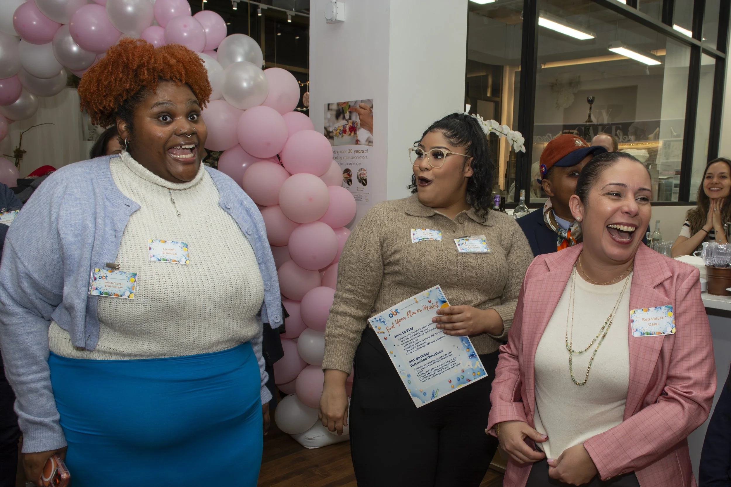 Three women are smiling and laughing at a celebration event with pink and white balloons in the background. They are wearing casual and semi-formal clothing, with name tags on their shirts.