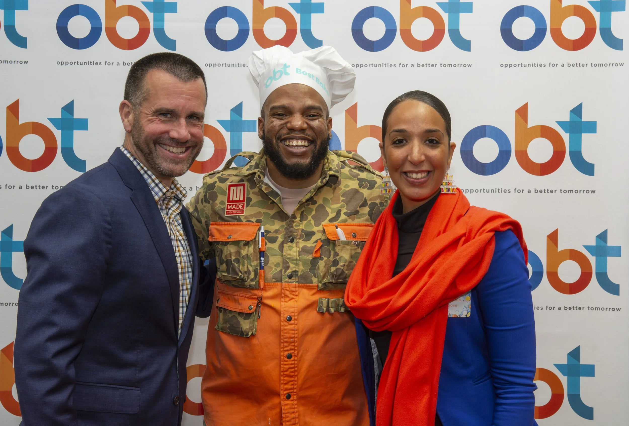 Three smiling people standing in front of a backdrop with the 'obt' logo and slogan. From left to right: a man in a navy blazer and checkered shirt, a man in camouflage and orange workwear with a chef's hat, and a woman in a blue dress with an orange