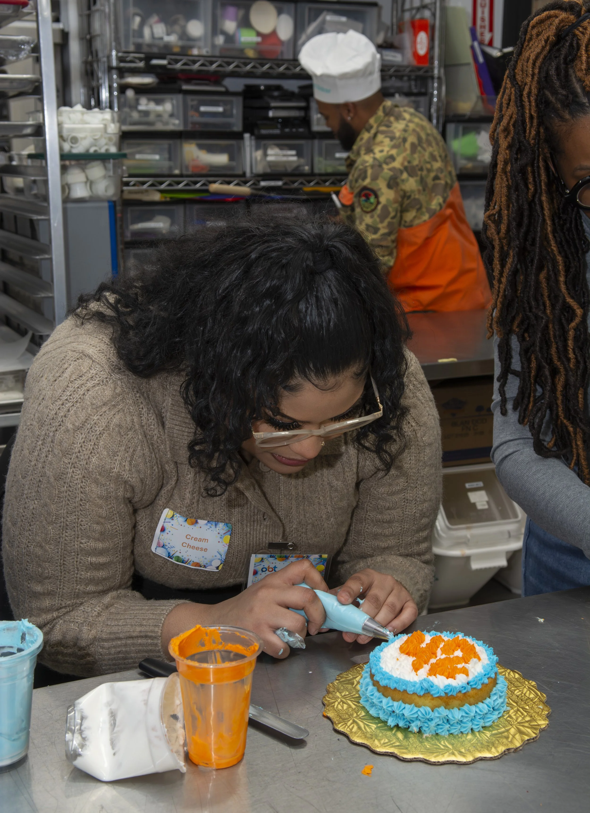 A woman with curly dark hair and glasses decorating a cake with orange and white frosting, in a kitchen or bakery setting.
