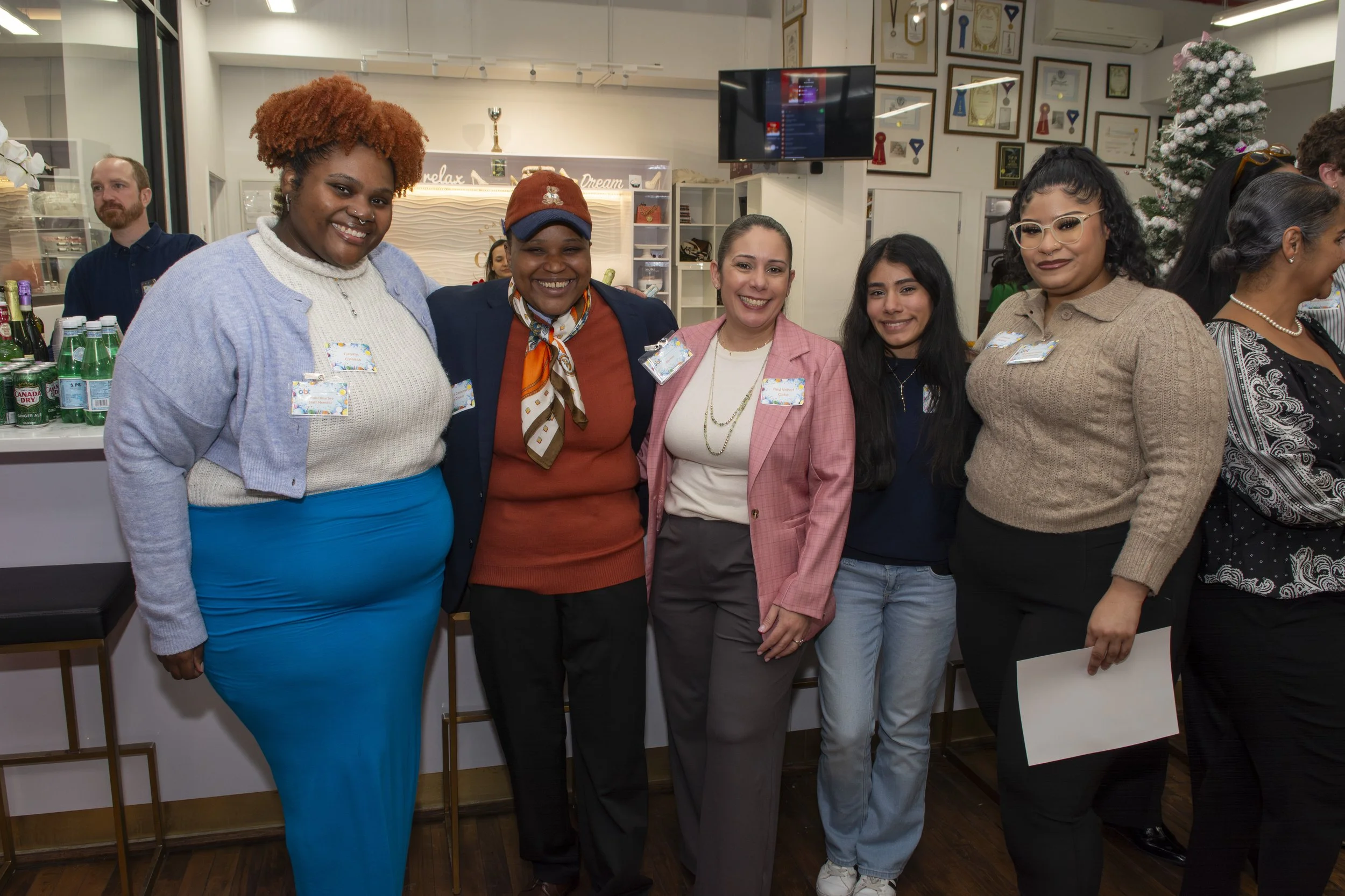 Group of six women standing together indoors, smiling for the camera at a celebration or gathering, with framed certificates and a Christmas tree in the background.