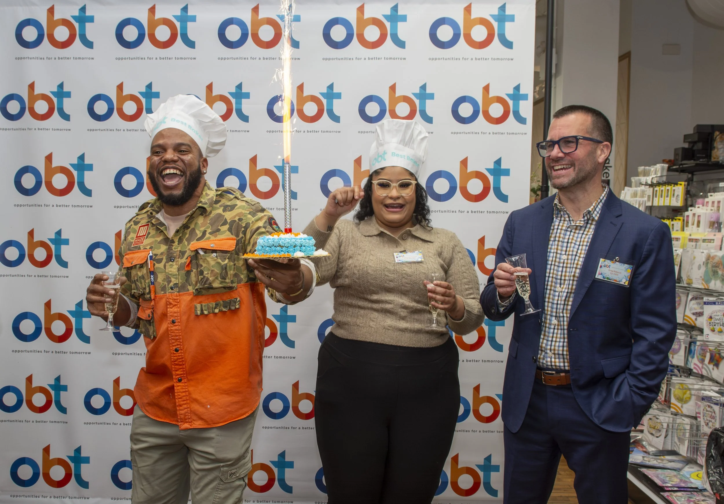 Three people celebrating a birthday at an event, standing in front of a backdrop with the OBOT logo. The person on the left holds a glass of bubbly and a birthday cake with a sparkler. The woman in the middle is wearing a chef's hat and glasses, smil