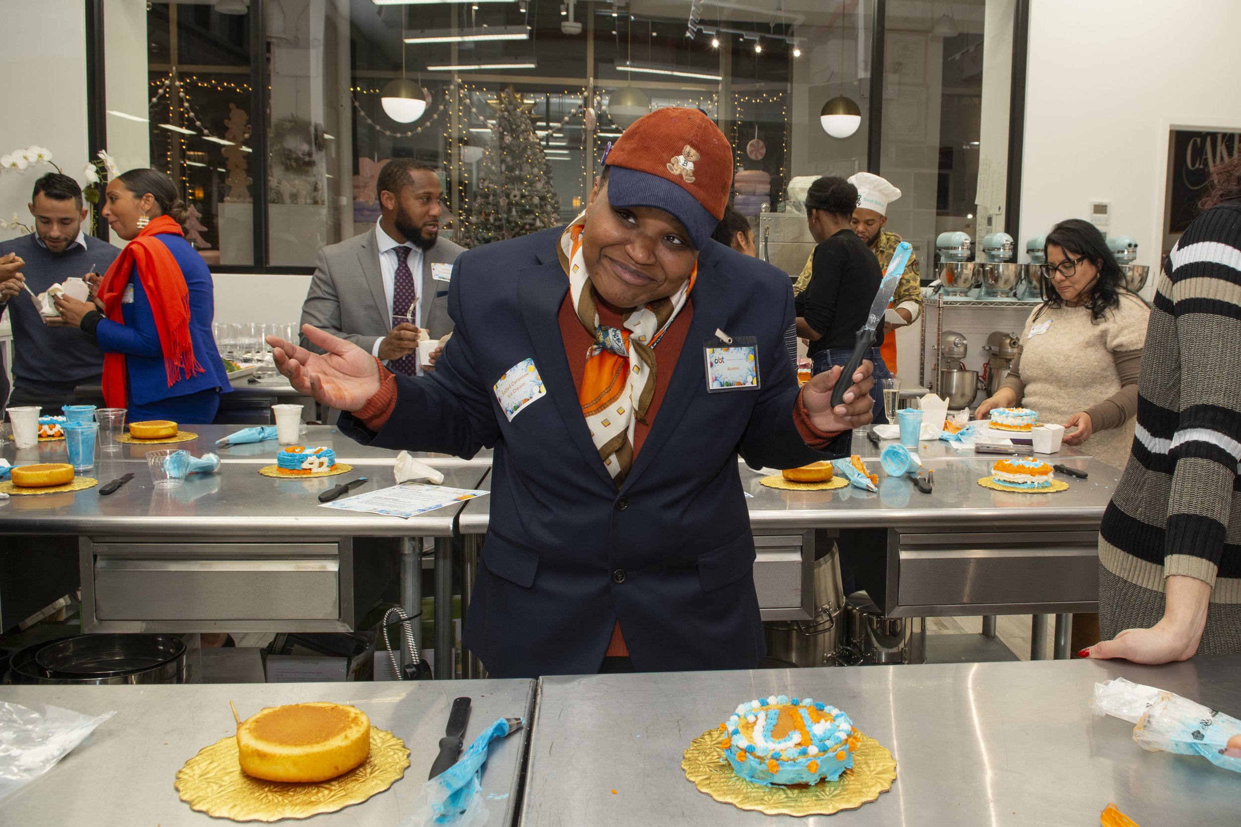 A woman in a navy blazer and a colorful scarf from the thumbnail is smiling and gesturing with her hands while holding a cake serving utensil during a cake decorating event in a kitchen. There are decorated cakes, knives, and icing on the table, and 