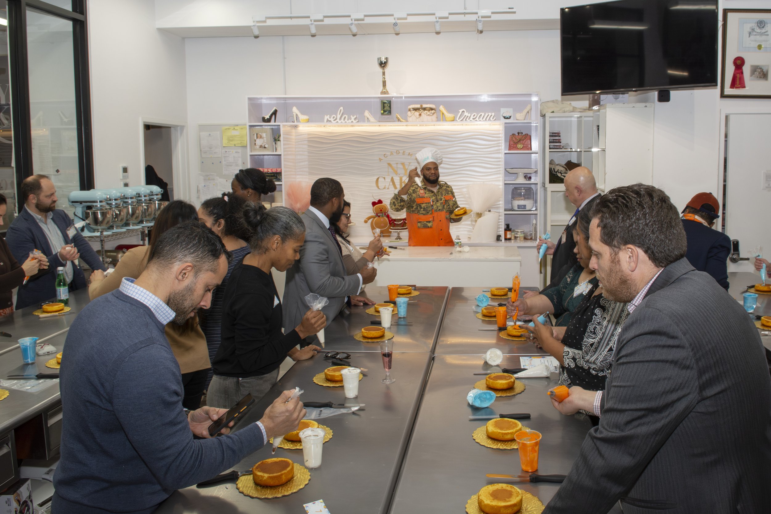 A group of people seated at a long table with cakes and decorating supplies, watching a chef in an orange apron and chef's hat demonstrate how to decorate cakes in a bakery setting.