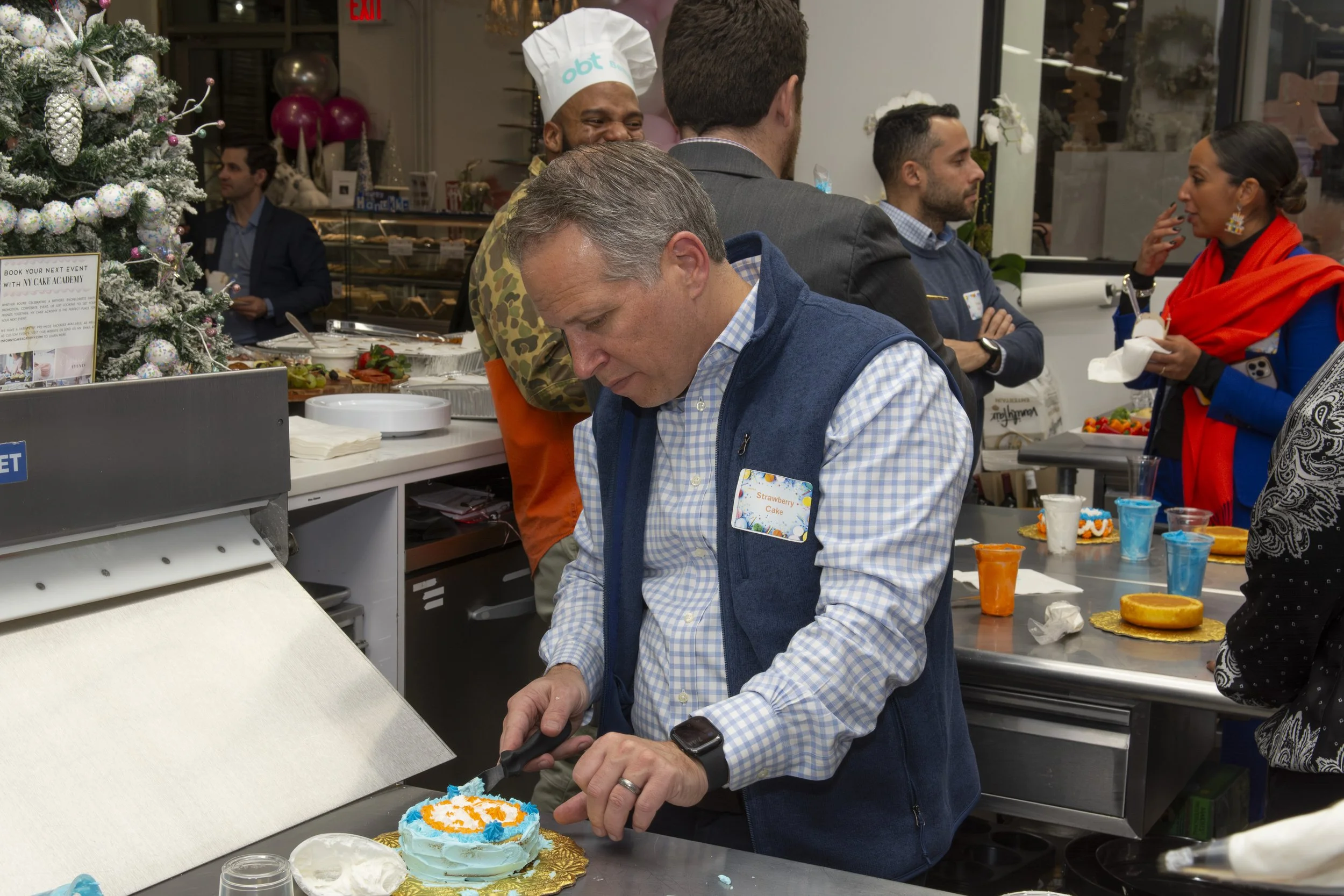 A man with a name tag that says 'Strawberry Cake' is decorating a birthday cake with blue and orange icing during a celebration in a bakery or café. Other people are gathered around, with some in conversation and some observing. There are holiday dec