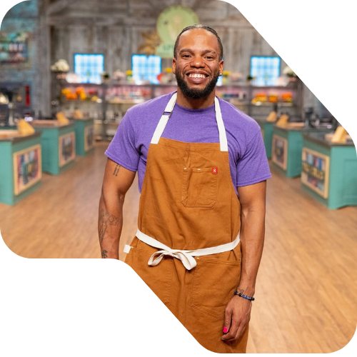 Baker and digital marketer kareem youngblood Smiling in a purple shirt and brown apron standing inside a colorful store or market with shelves and displays. on the set of spring baking championship