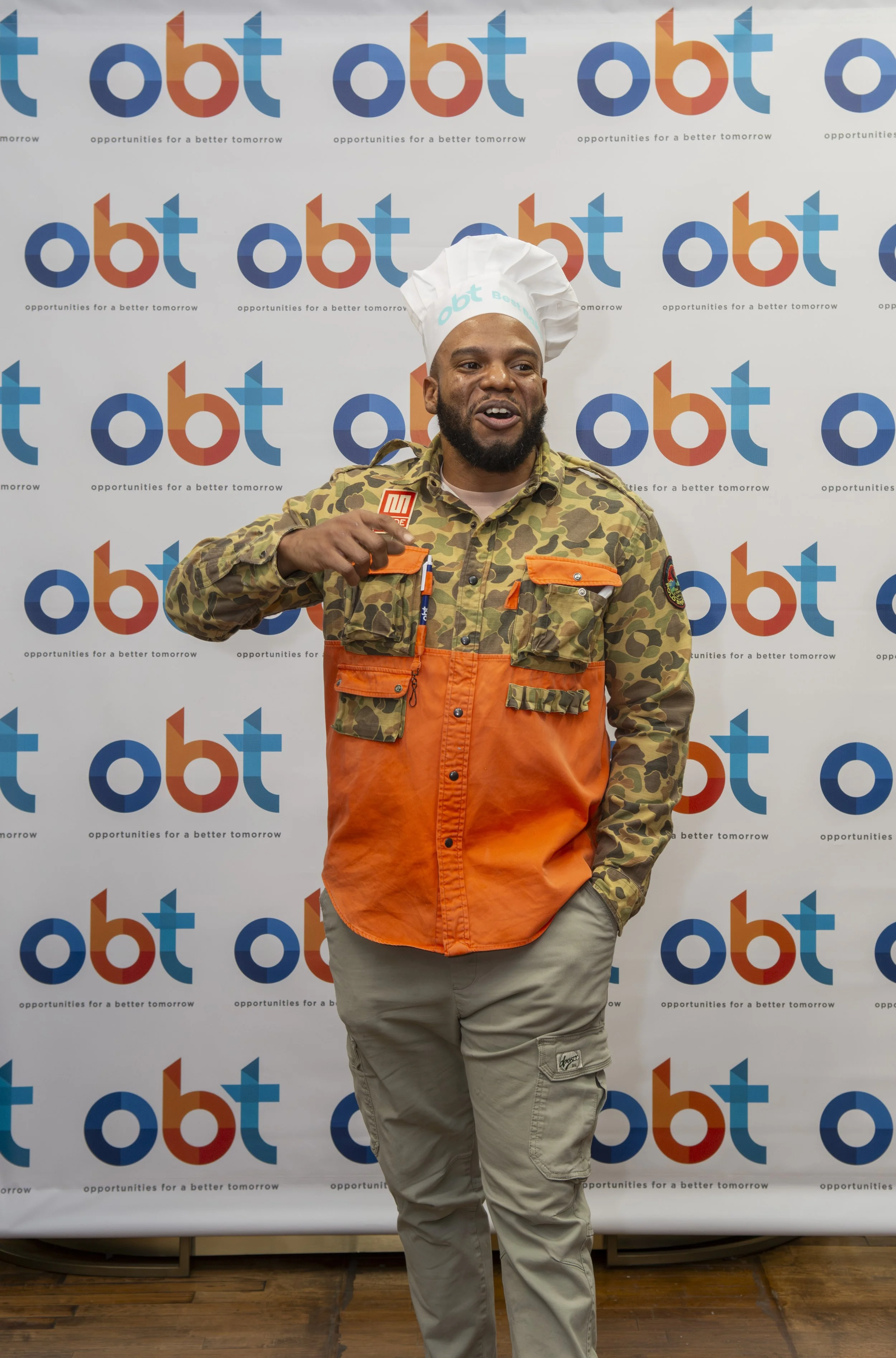Man in camouflage and orange work shirt wearing a chef hat, standing in front of a backdrop with 'OBT' logos, smiling and pointing at his chest.
