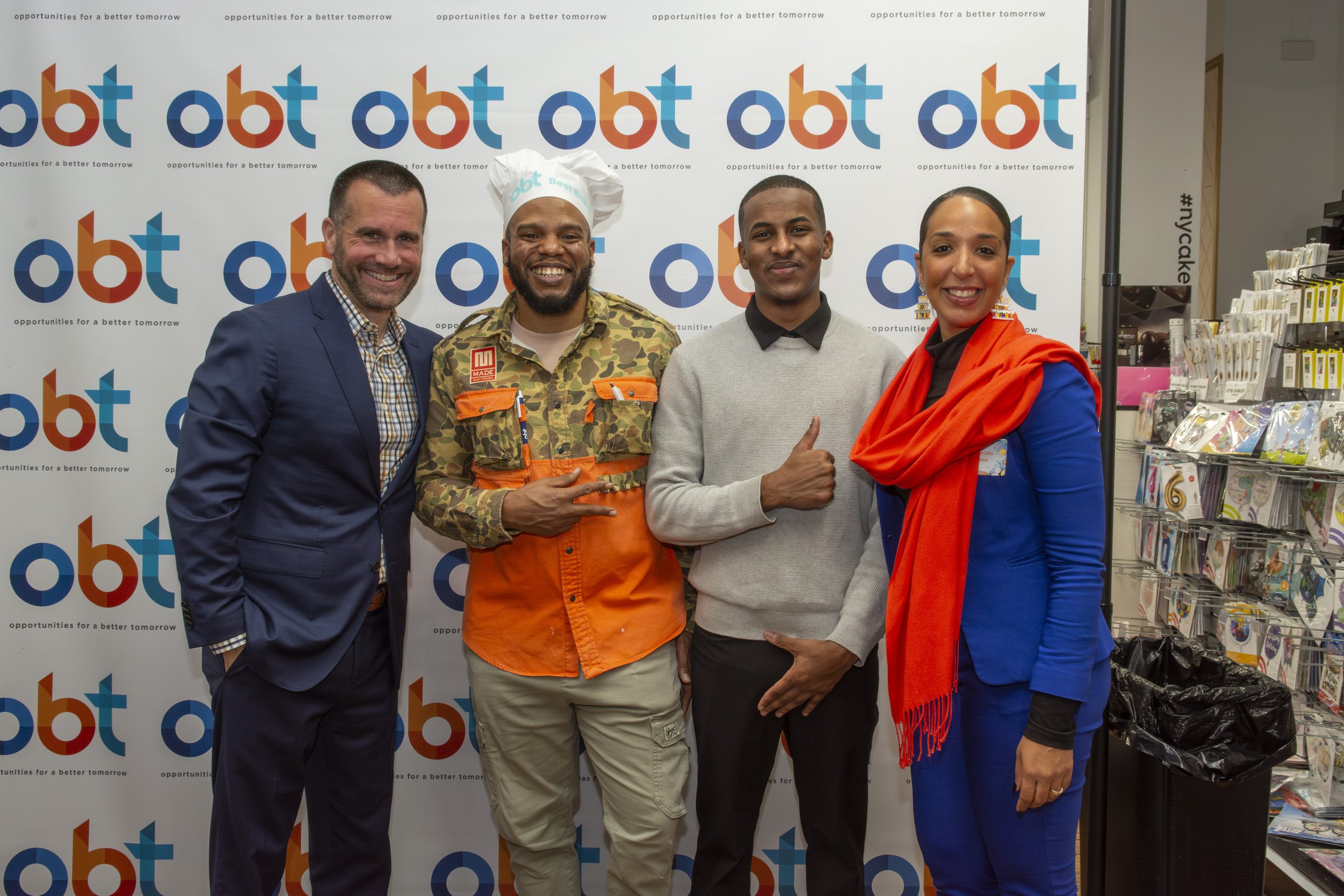 Four people standing in front of a step and repeat banner with the logo 'obt' and the slogan 'opportunities for a better tomorrow.' They are smiling and posing for a photo, with some making hand gestures. The background includes shelves with various 