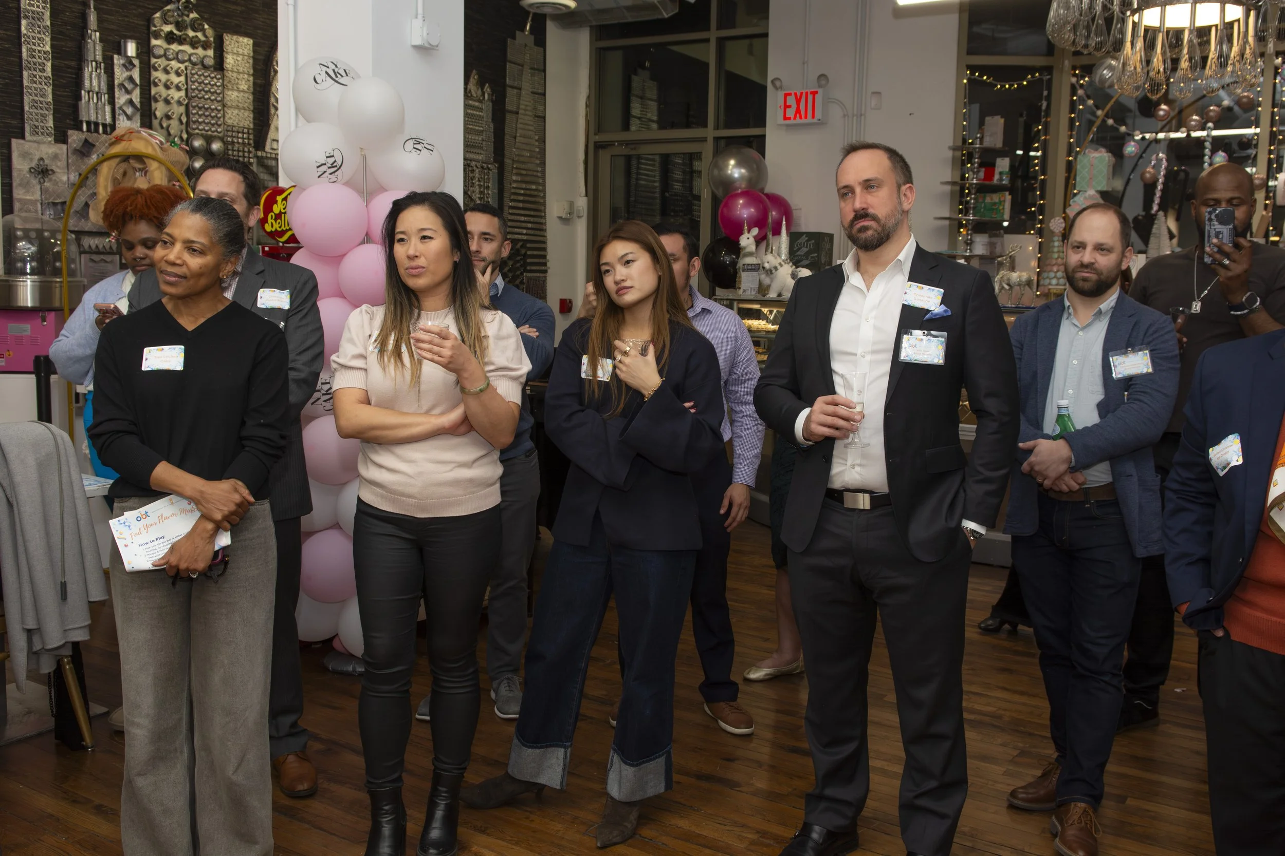 Group of diverse adults at a social gathering, listening attentively, in a decorated indoor space with pink and white balloons and string lights.