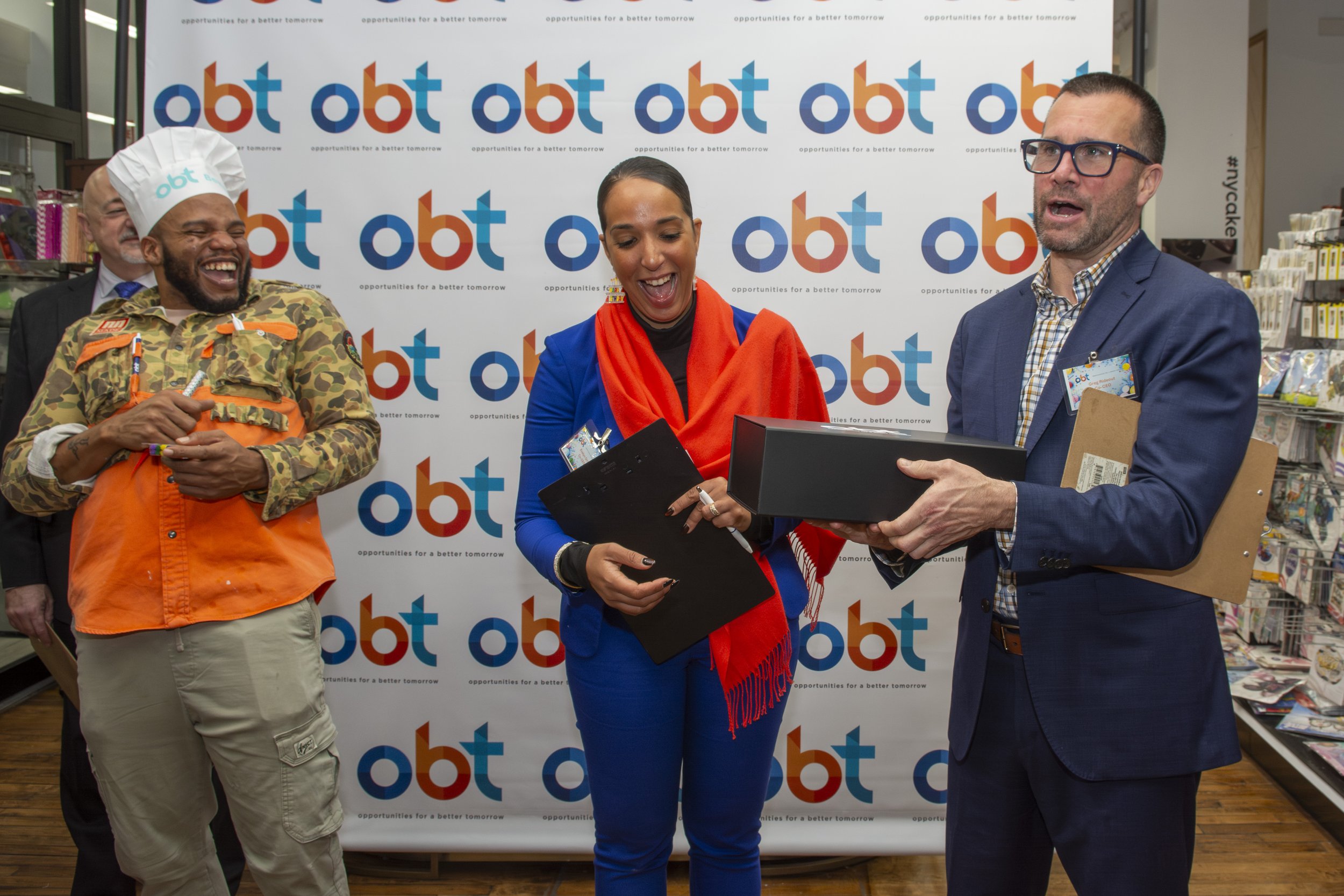 Four people at an event standing in front of a backdrop with the 'obt' logo. One man is wearing a camouflage jacket and orange vest, smiling, holding a pen. The woman in the center is holding a clipboard and smiling. The man on the right, wearing gla