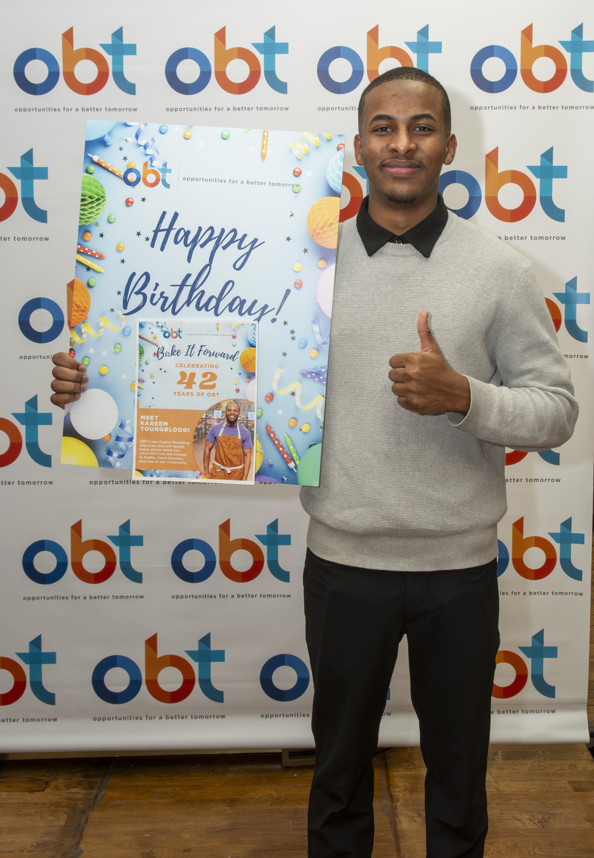 A man standing in front of a backdrop with 'OBT' logos, holding a birthday sign that says 'Happy Birthday!' with colorful decorations, and giving a thumbs-up gesture.