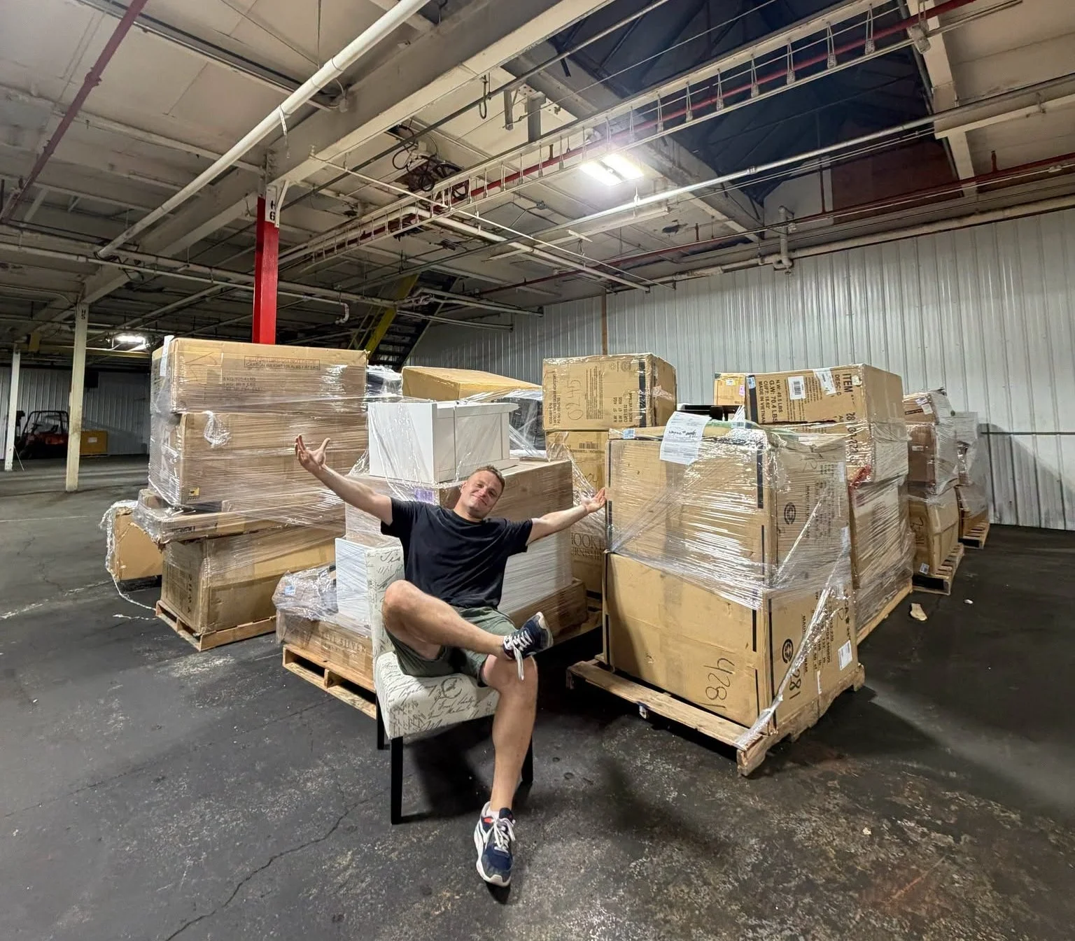 A man sitting on a chair in a warehouse surrounded by large stacks of wrapped boxes and pallets, with one leg propped up and arms open in a gestures of enthusiasm.