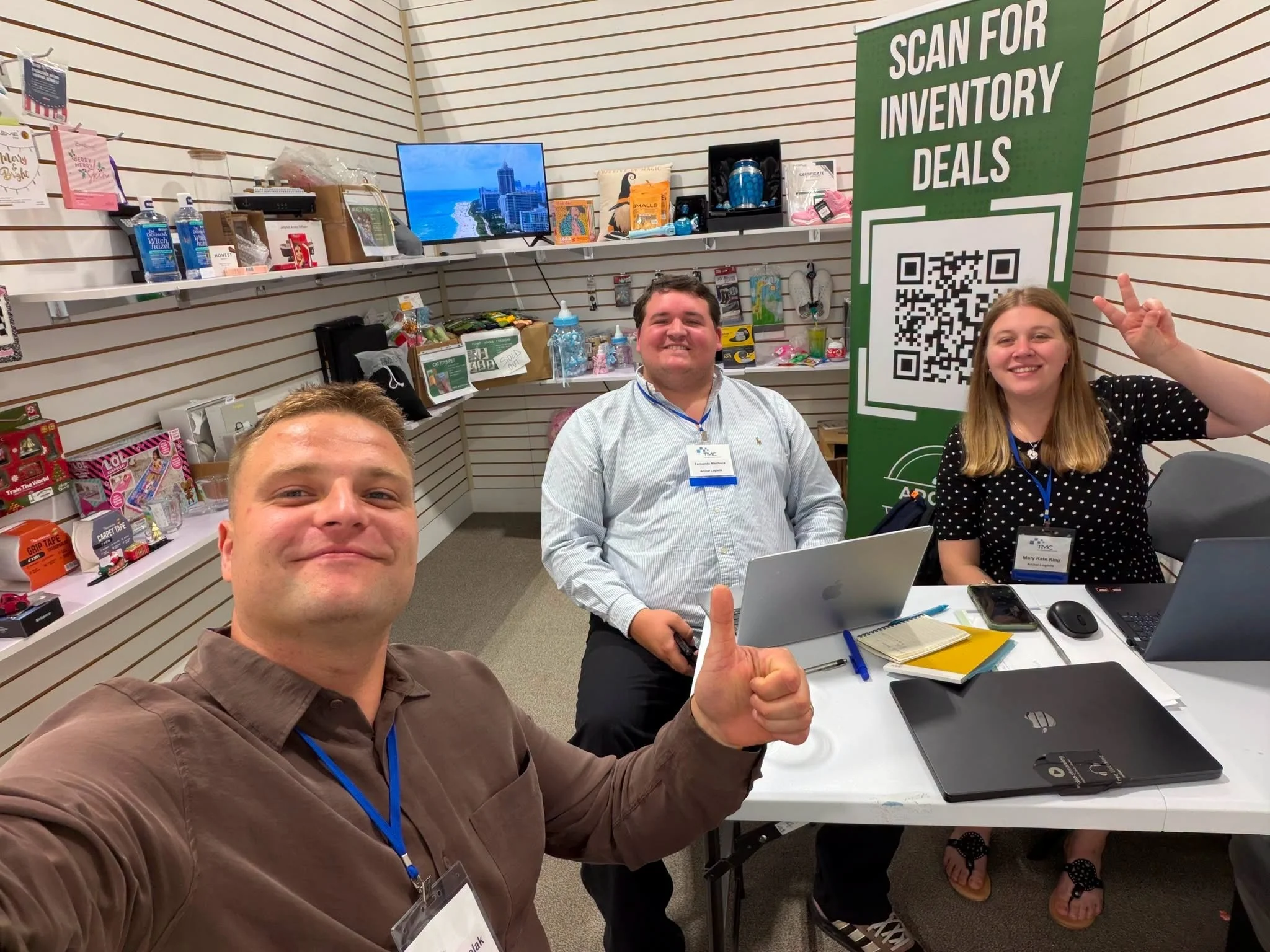 Three people smiling and posing for a selfie at a conference or meeting, with one giving a thumbs-up. The setting has a display shelf with various items and a large QR code sign that reads "Scan for Inventory Deals." The table in front of them has laptops, notebooks, and pens. There are also other attendees visible in the background.