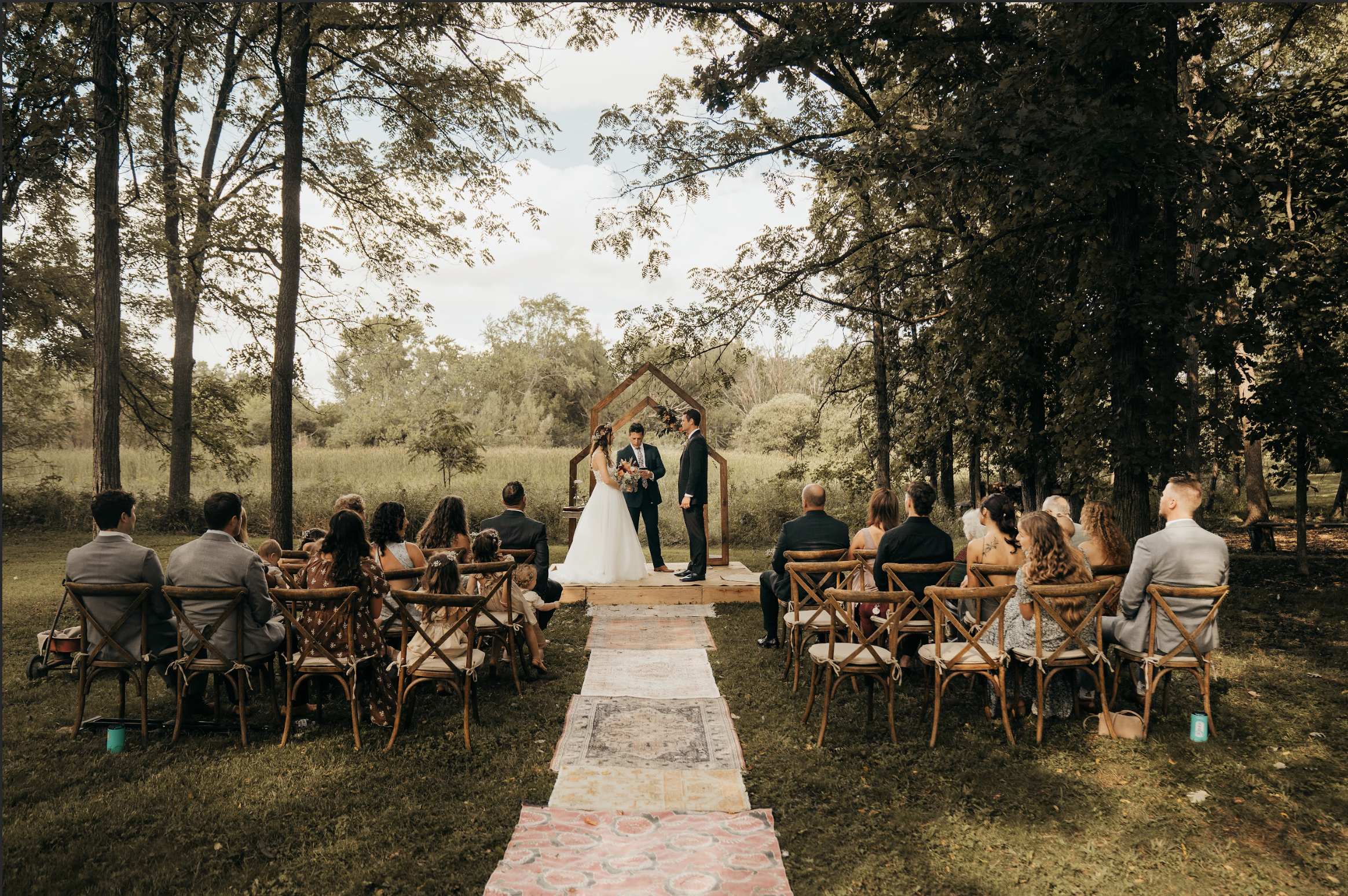A wedding ceremony taking place outdoors in a park or garden, with a bride and groom standing under a wooden arch, surrounded by seated guests, trees, and greenery.