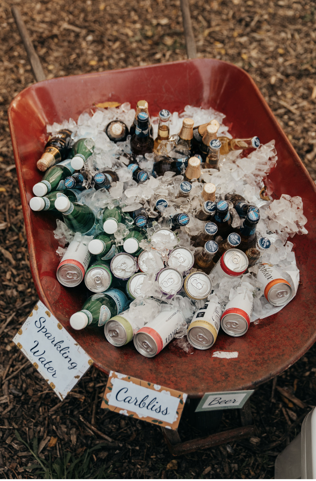 A red wheelbarrow filled with ice containing various bottles of beer and sparkling water, with handwritten signs for 'Sparkling Water', 'Cabiliss', and 'Beer' placed around it.