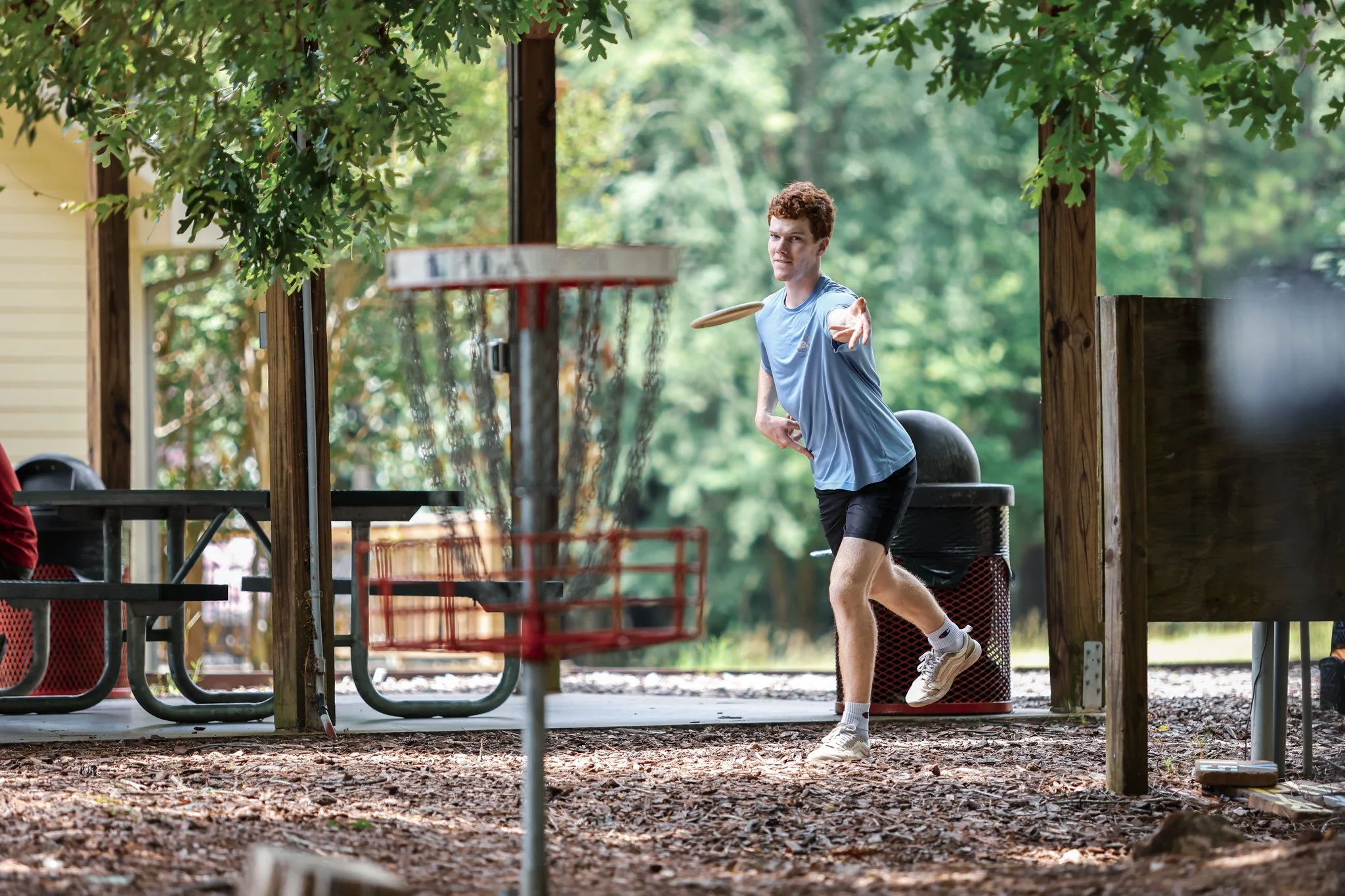 A young man playing disc golf outdoors, throwing a disc towards a basket, under green trees with a picnic table and trash can in the background.