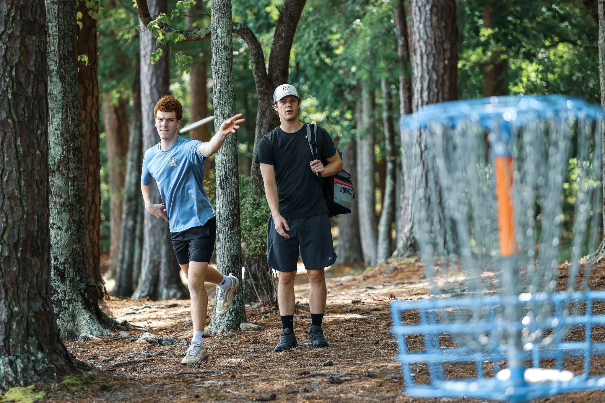 Two young men playing disc golf in a wooded forest, one tossing a disc towards a blue disc golf basket.