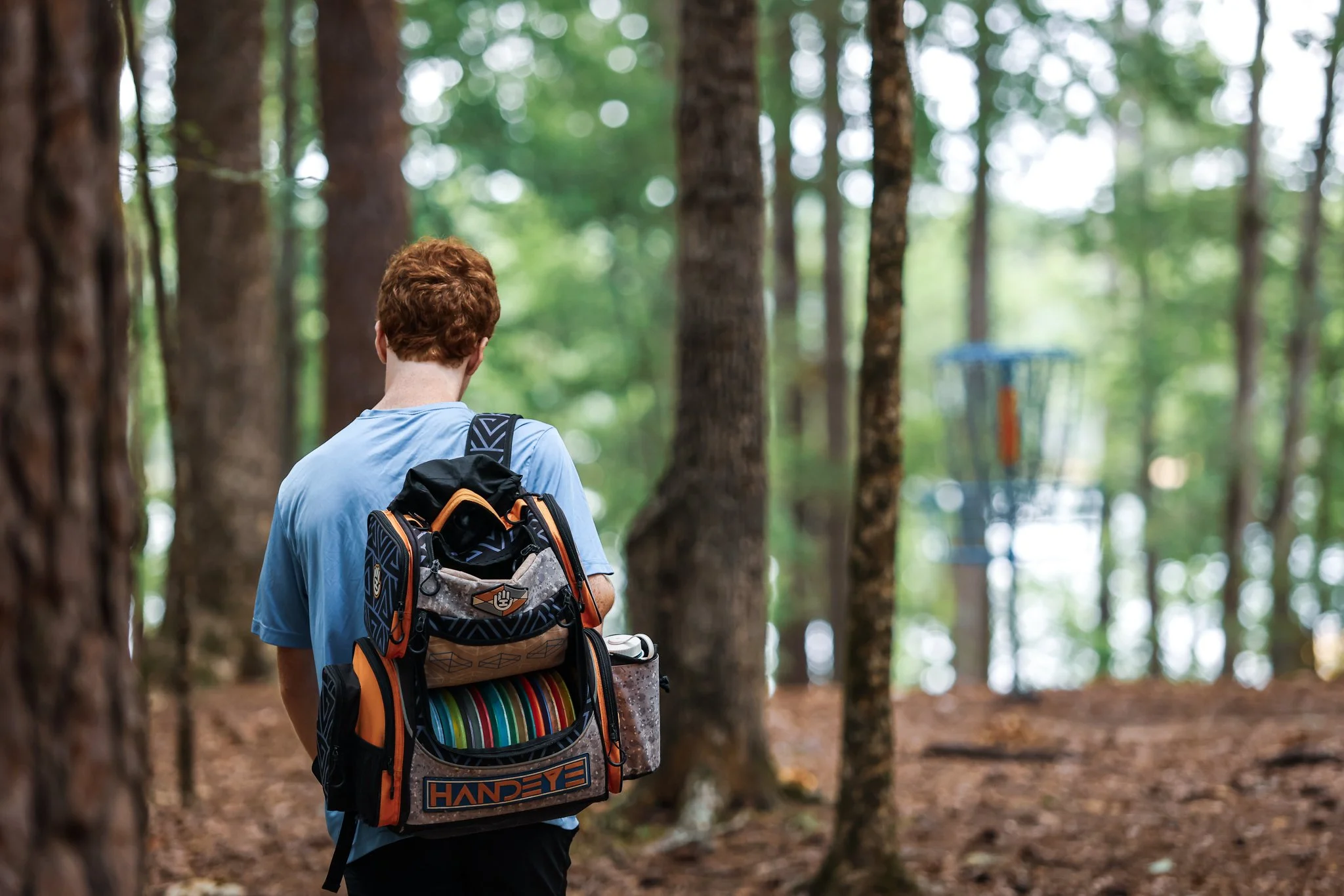 A person with red hair wearing a blue shirt and a backpack, heading through a forest towards a disc golf basket in the distance.