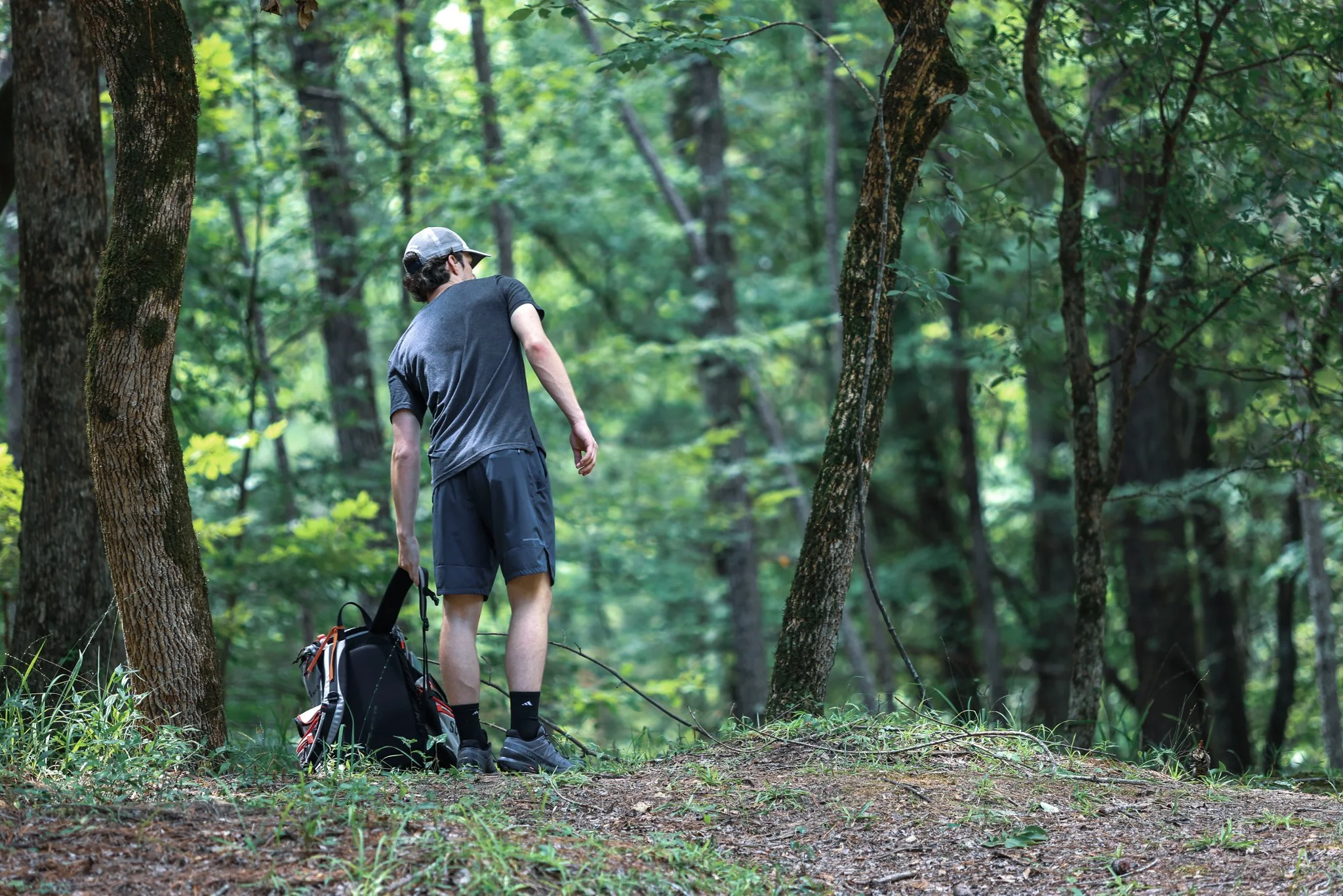 A man wearing a gray cap and black athletic clothing stands in a green forest, holding a black backpack on the ground, looking at the trees and forest floor.