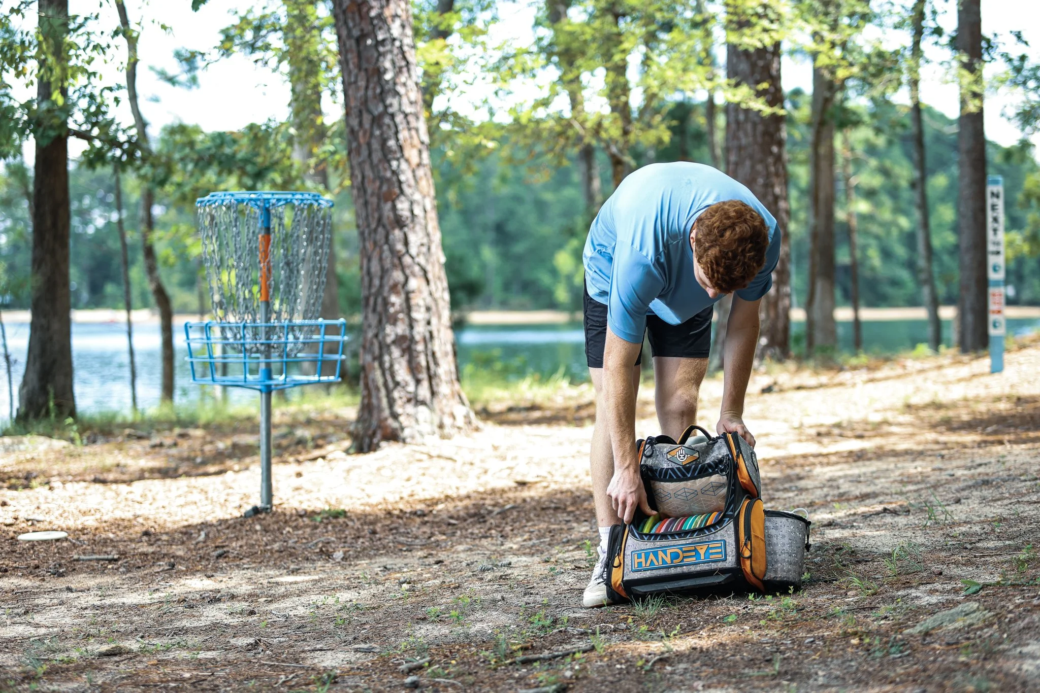 A man in a blue shirt and black shorts prepares for disc golf at a wooded park near a lake, with a disc golf basket visible nearby.