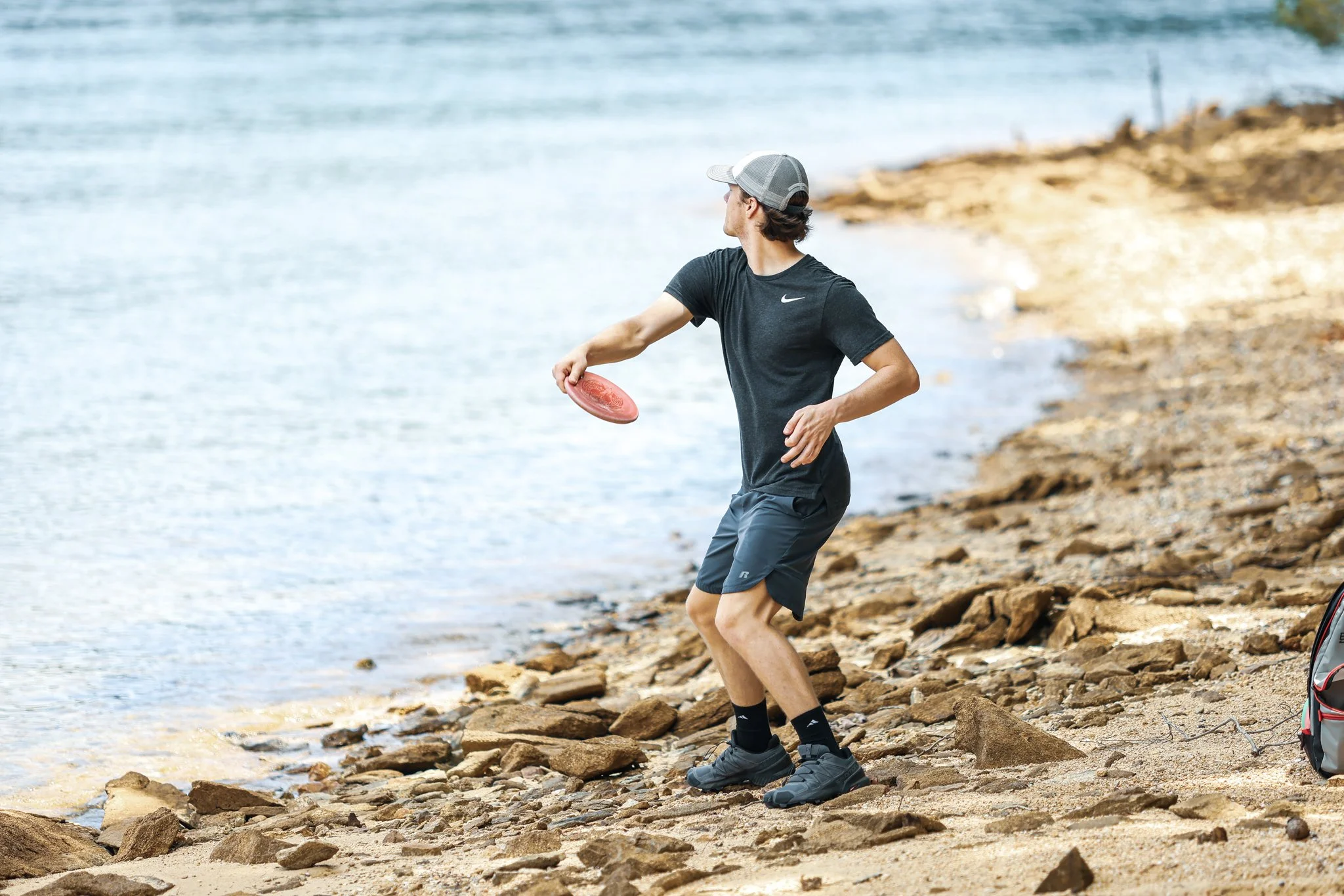 Man playing disc golf on rocky beach near water, wearing black athletic shirt, shorts, and gray cap.