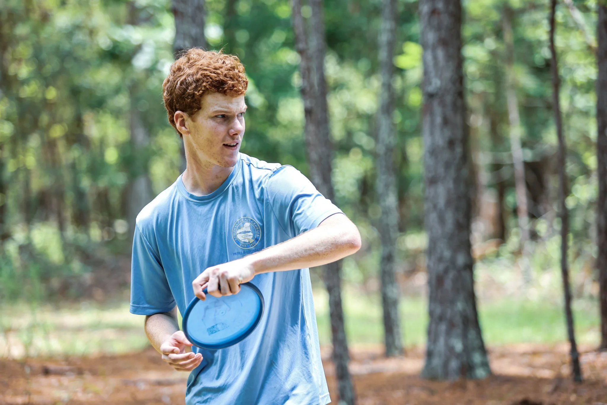 A young man with red hair and freckles in a blue T-shirt standing outdoors in a wooded area, holding a frisbee and looking to the left.