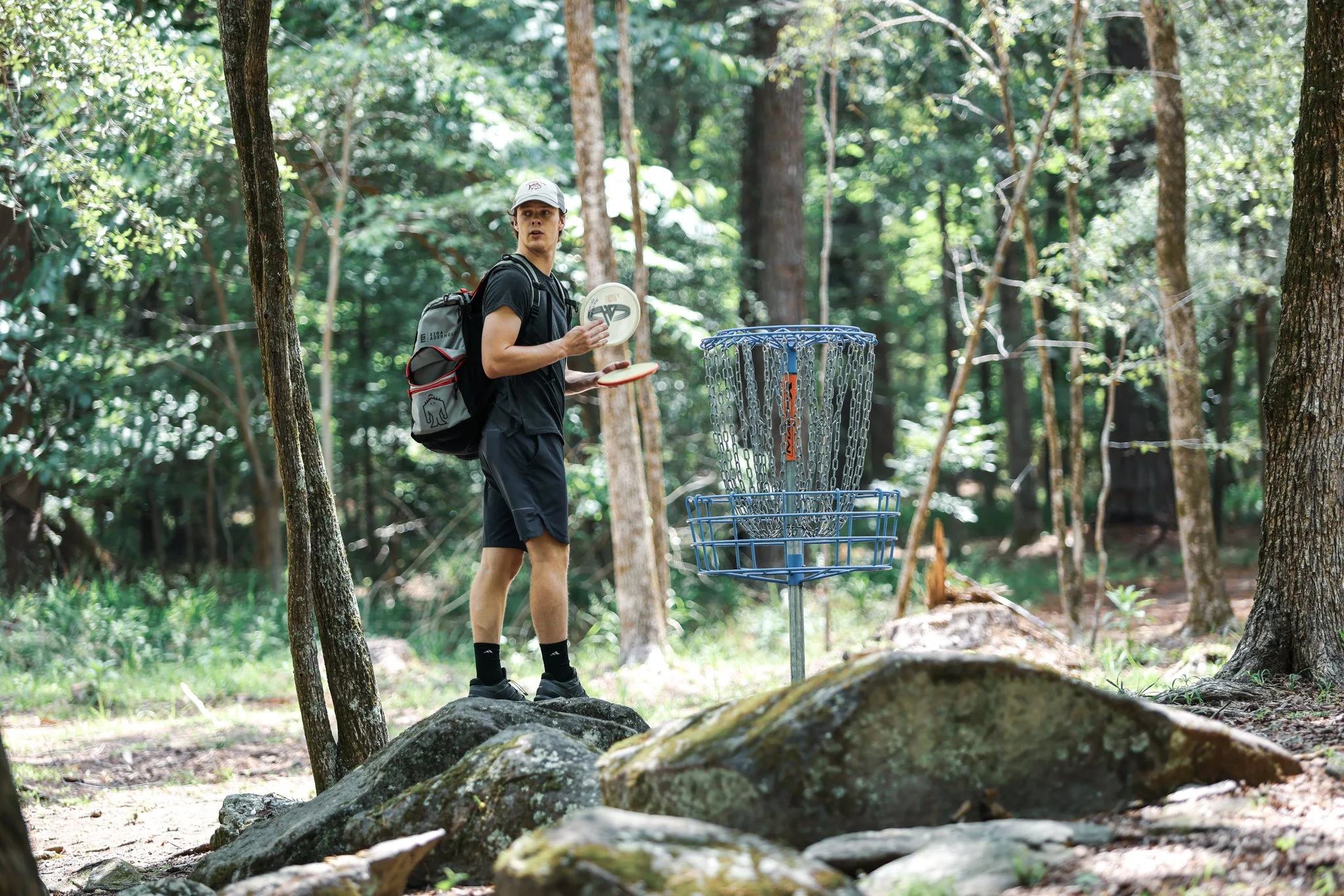 A person standing in a wooded area about to throw a disc towards a disc golf basket.