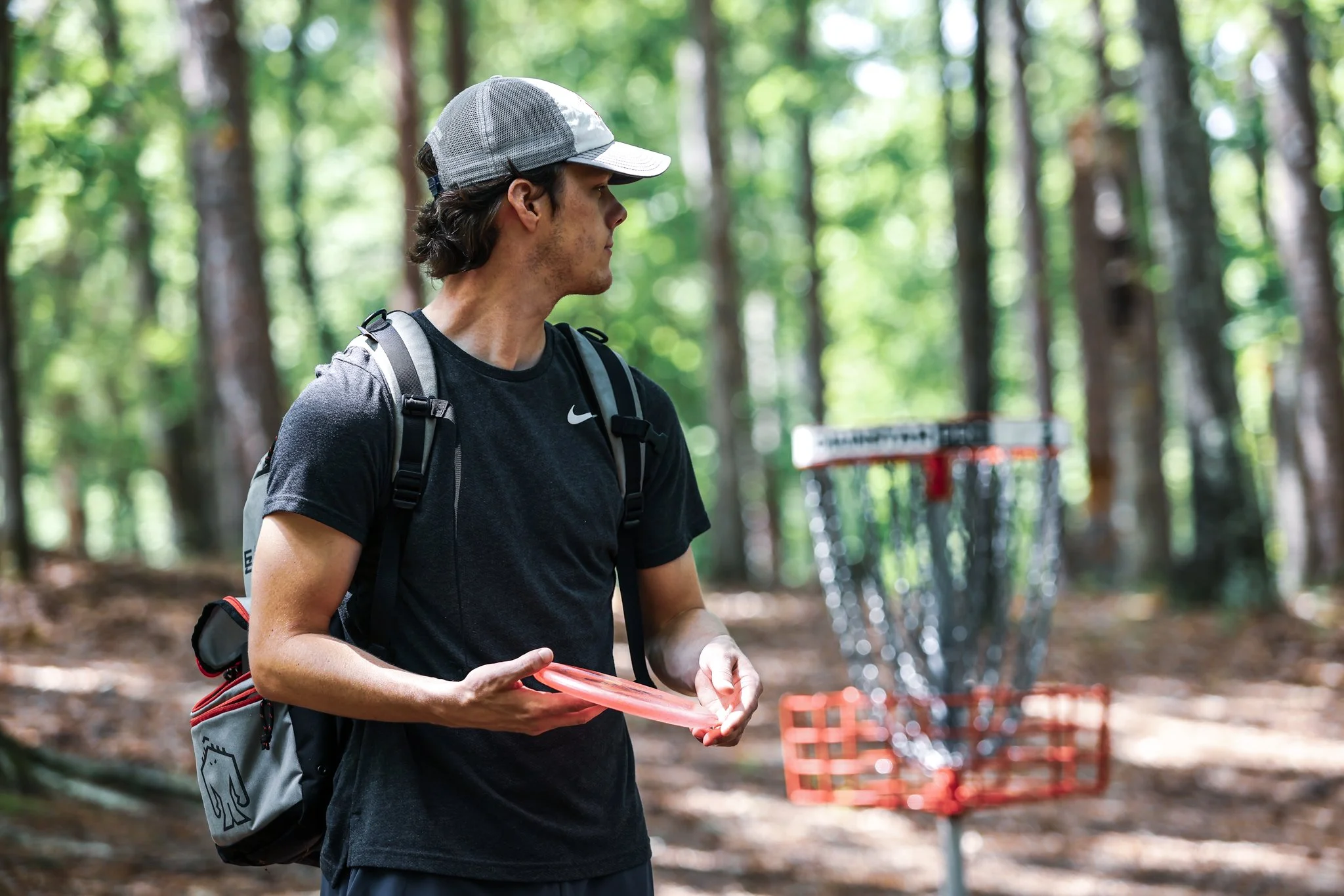 A young man with long brown hair, wearing a black T-shirt, gray baseball cap, and backpack, stands in a wooded area holding a disc golf disc, with a disc golf basket in the background.
